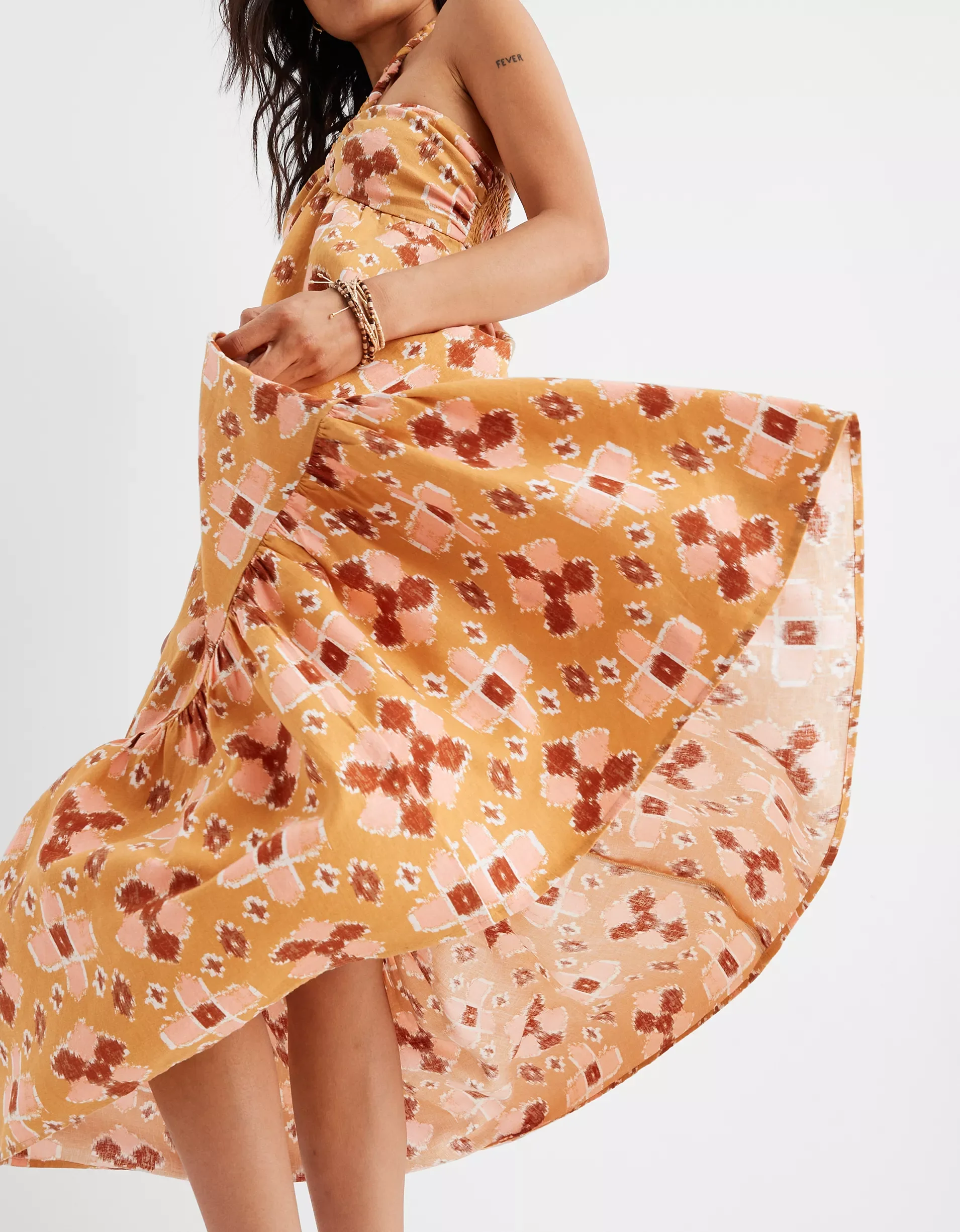 A woman wearing a flowy, floral-patterned dress in shades of orange, pink, and red holding up the skirt to reveal her legs against a plain white background.