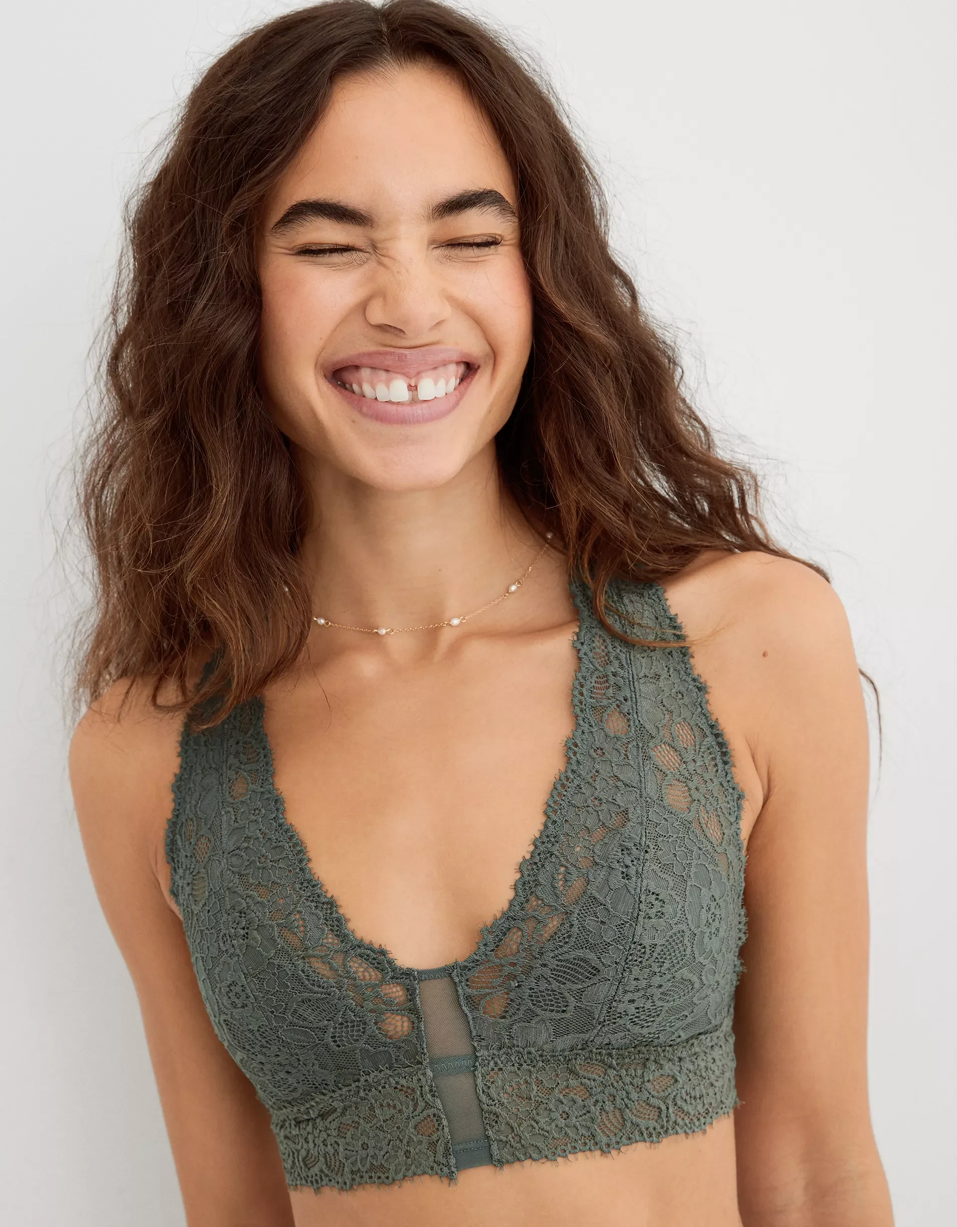 Young woman with long, curly brown hair smiling, wearing a green lace bralette and a delicate necklace.