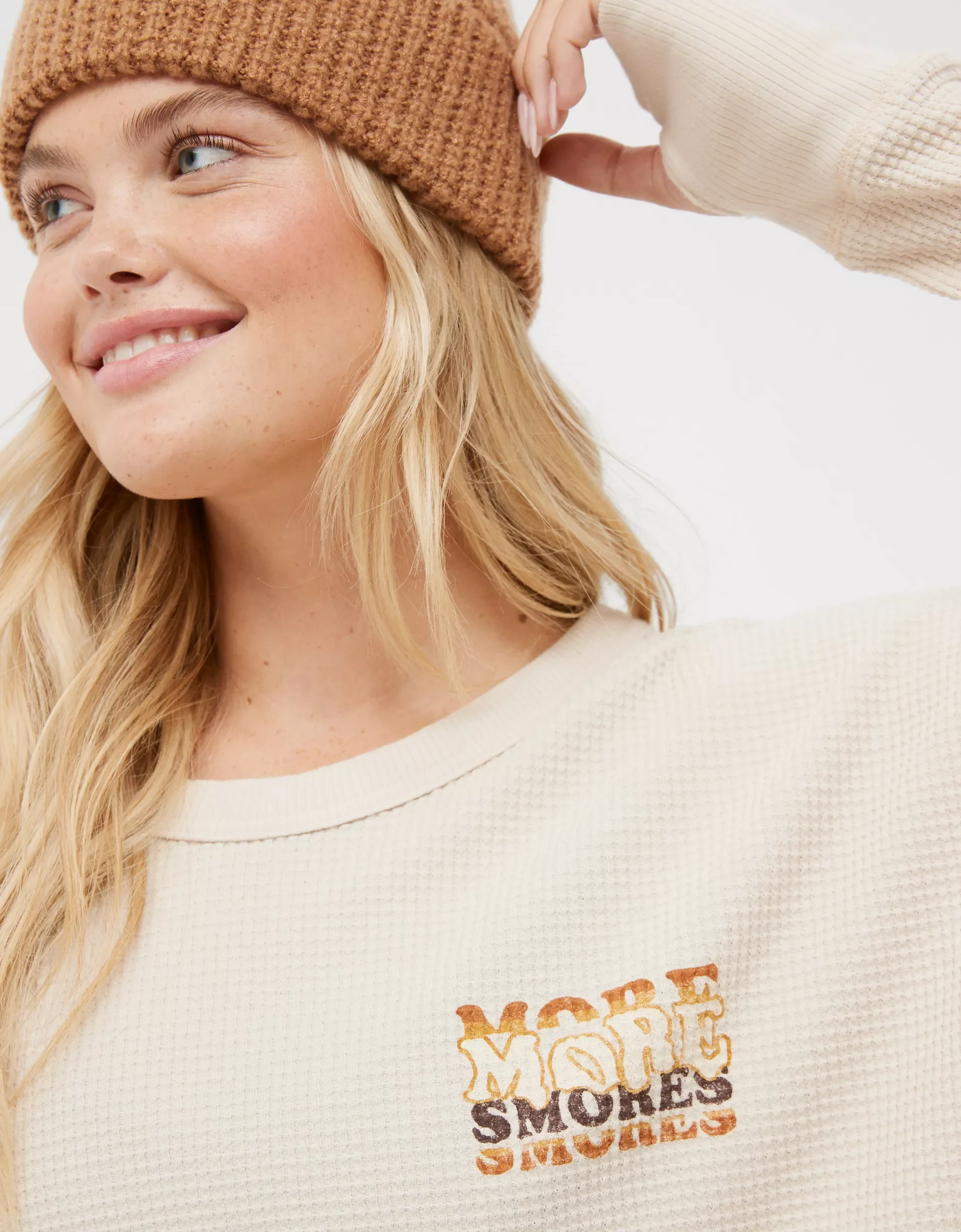 A young woman with long blonde hair smiling, wearing a beige sweatshirt with a 'More Smokes' graphic and a brown knit beanie, adjusting her beanie with her right hand.