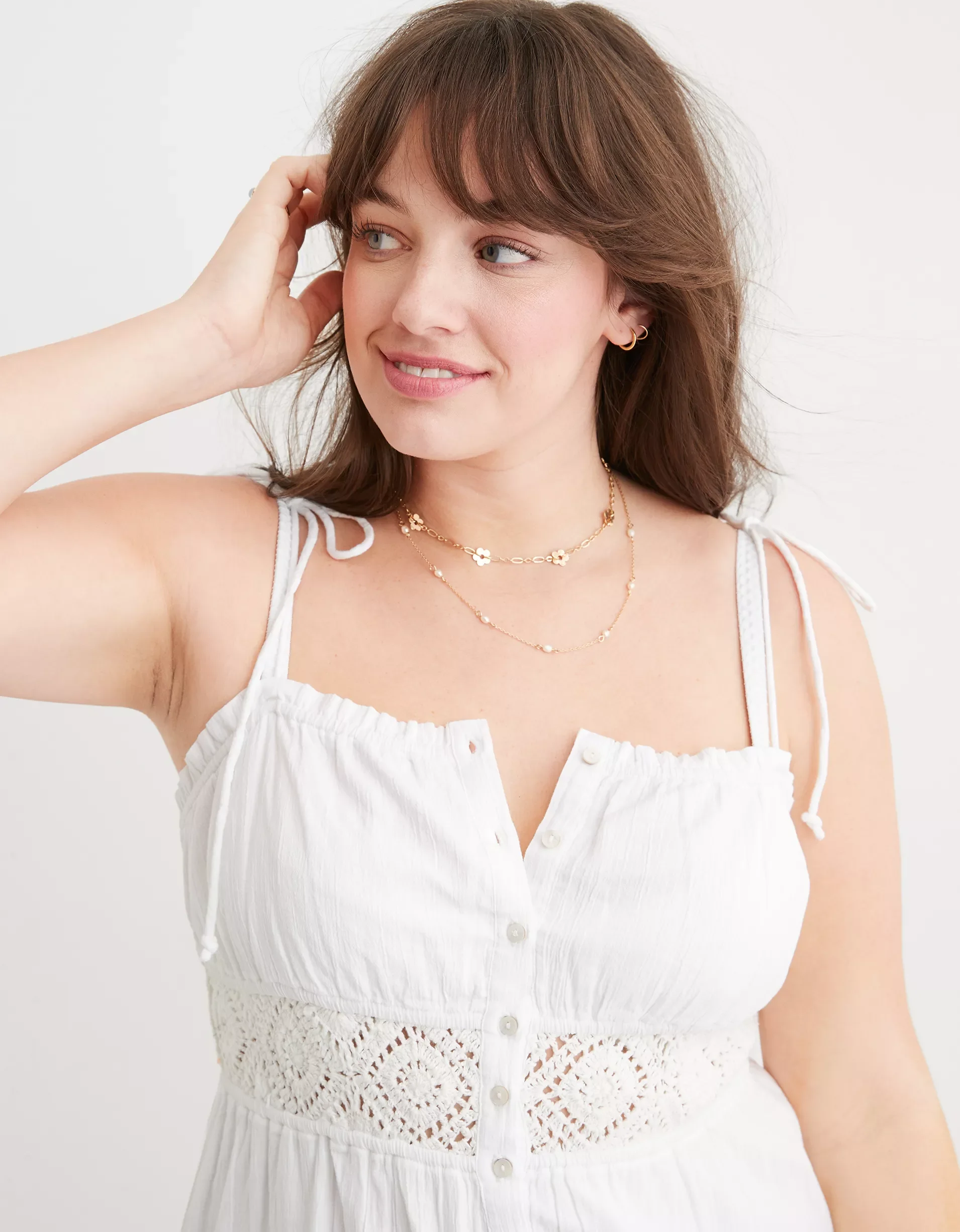 A young woman with brown hair wearing a white sleeveless dress, layered necklaces, and gold earrings, smiling softly against a plain white background.