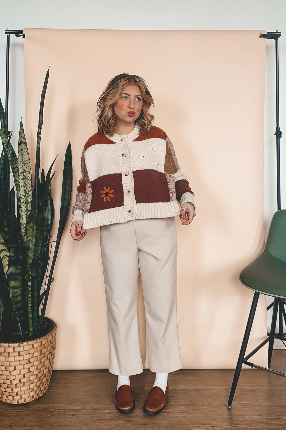 A woman with shoulder-length wavy hair, wearing a patchwork cardigan with floral embroidery, cream wide-leg pants, white socks, and brown loafers, standing in front of a beige backdrop with a green plant on her left and a green chair on her right.