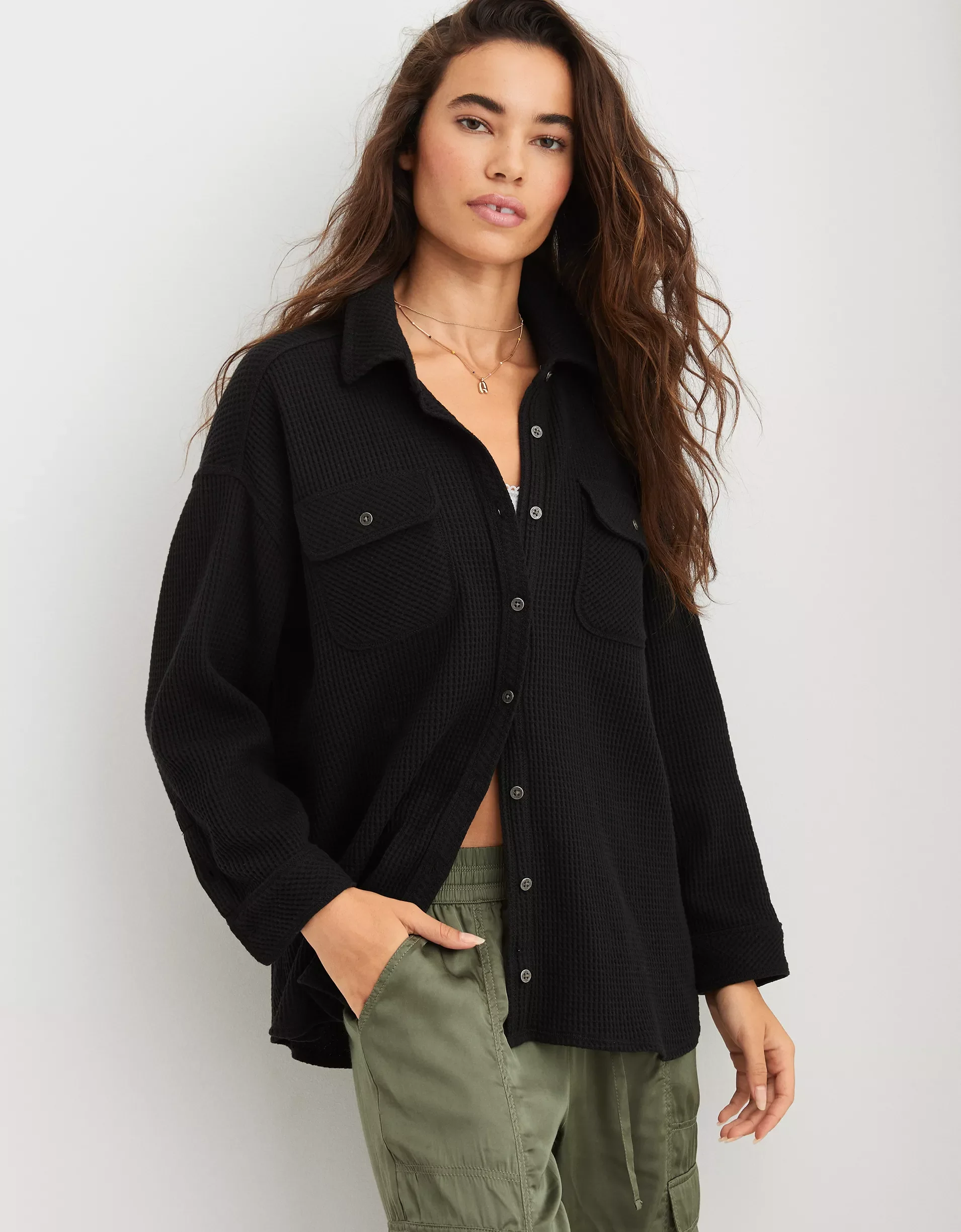 A woman with long wavy brown hair wearing a black loose-fitting knit button-up shirt, with olive green cargo pants and layered necklaces, standing against a plain white background.