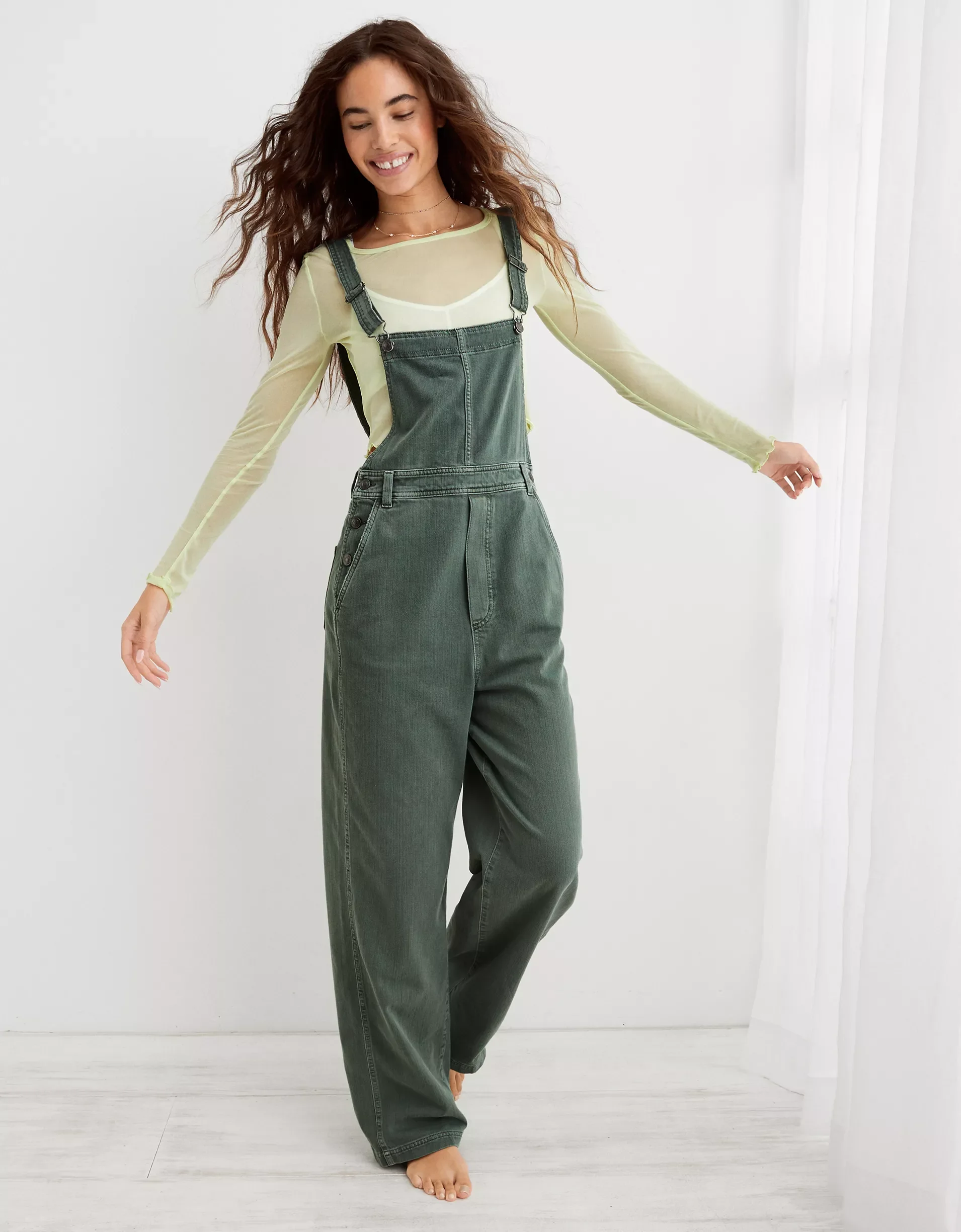 A woman smiling and twirling, wearing green overalls over a semi-transparent yellow long-sleeve top, barefoot on a white wooden floor near white curtains.