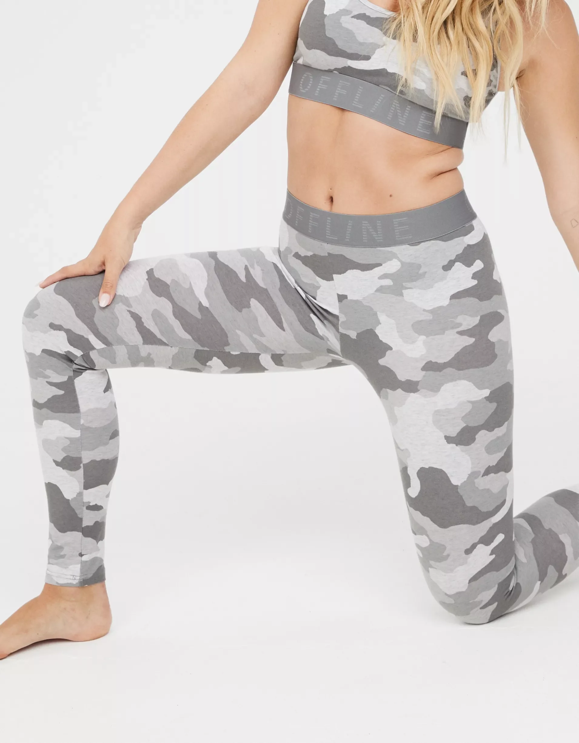 A woman in gray camouflage leggings and matching sports bra kneeling on one knee, with her left hand on her thigh and her right hand resting on her bent knee.