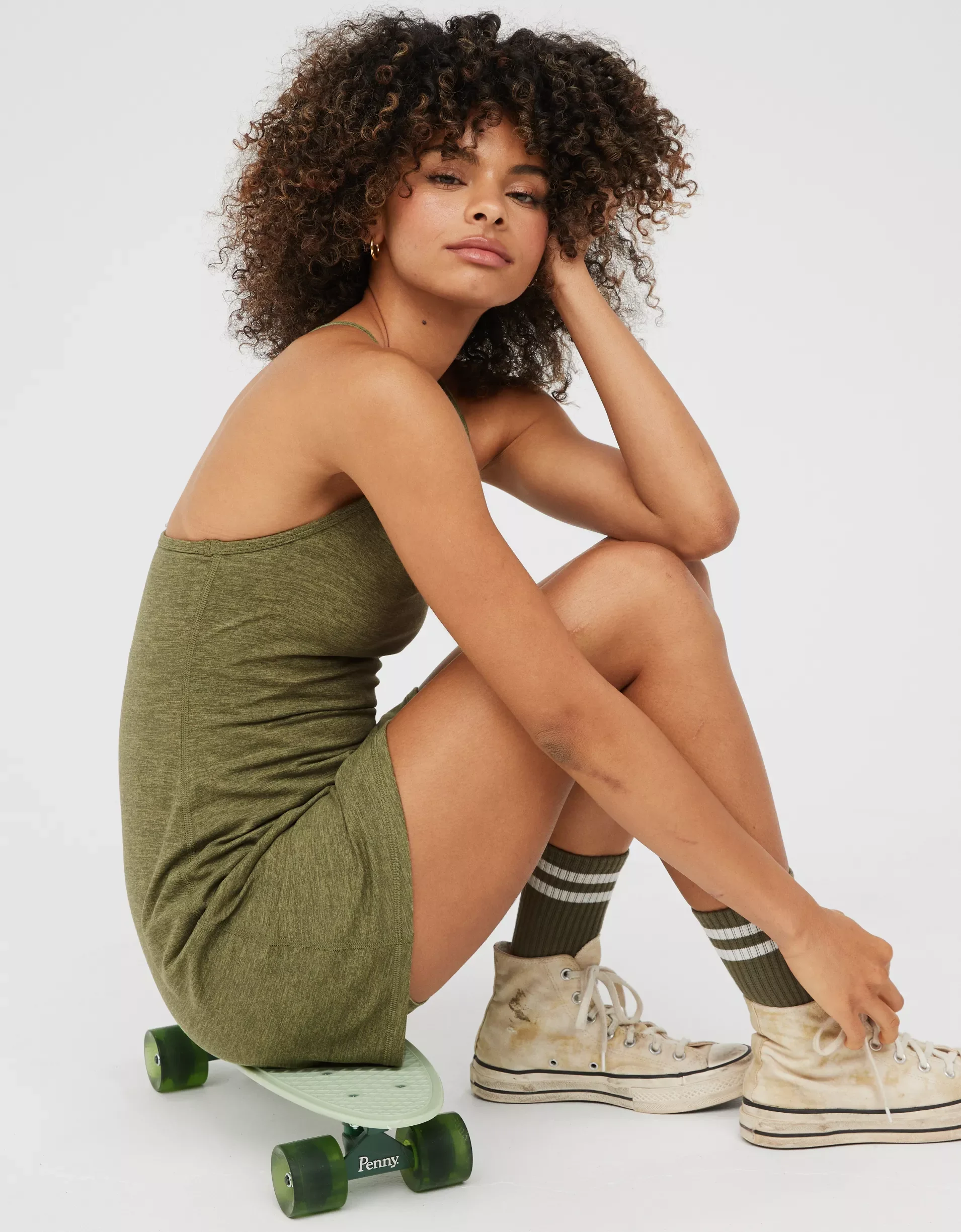 Young woman with curly hair in a green dress, sitting on a skateboard, wearing white high-top sneakers and striped socks, against a plain white background.