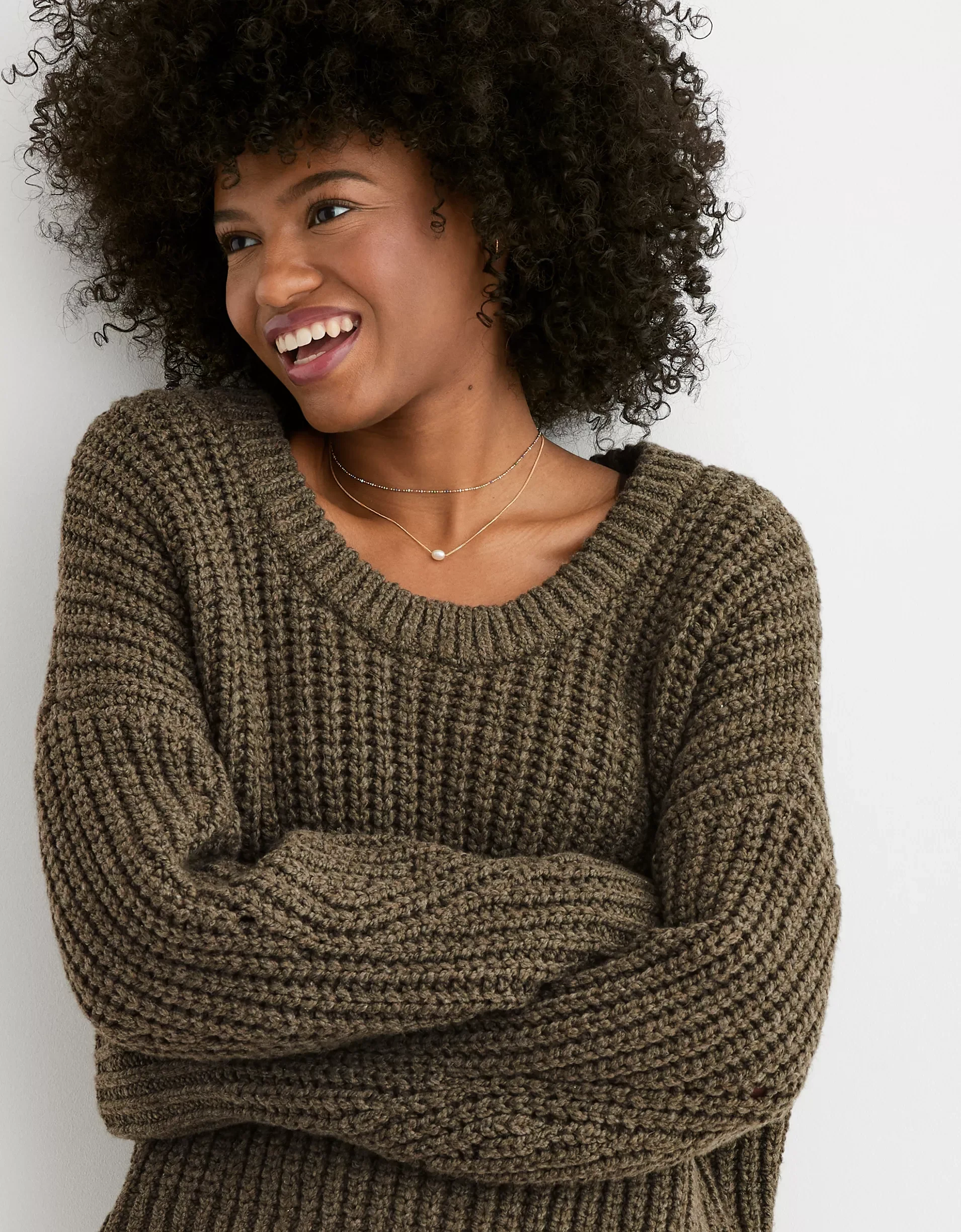 A woman with curly hair wearing a brown knit sweater, crossing her arms, smiling, against a plain white wall.