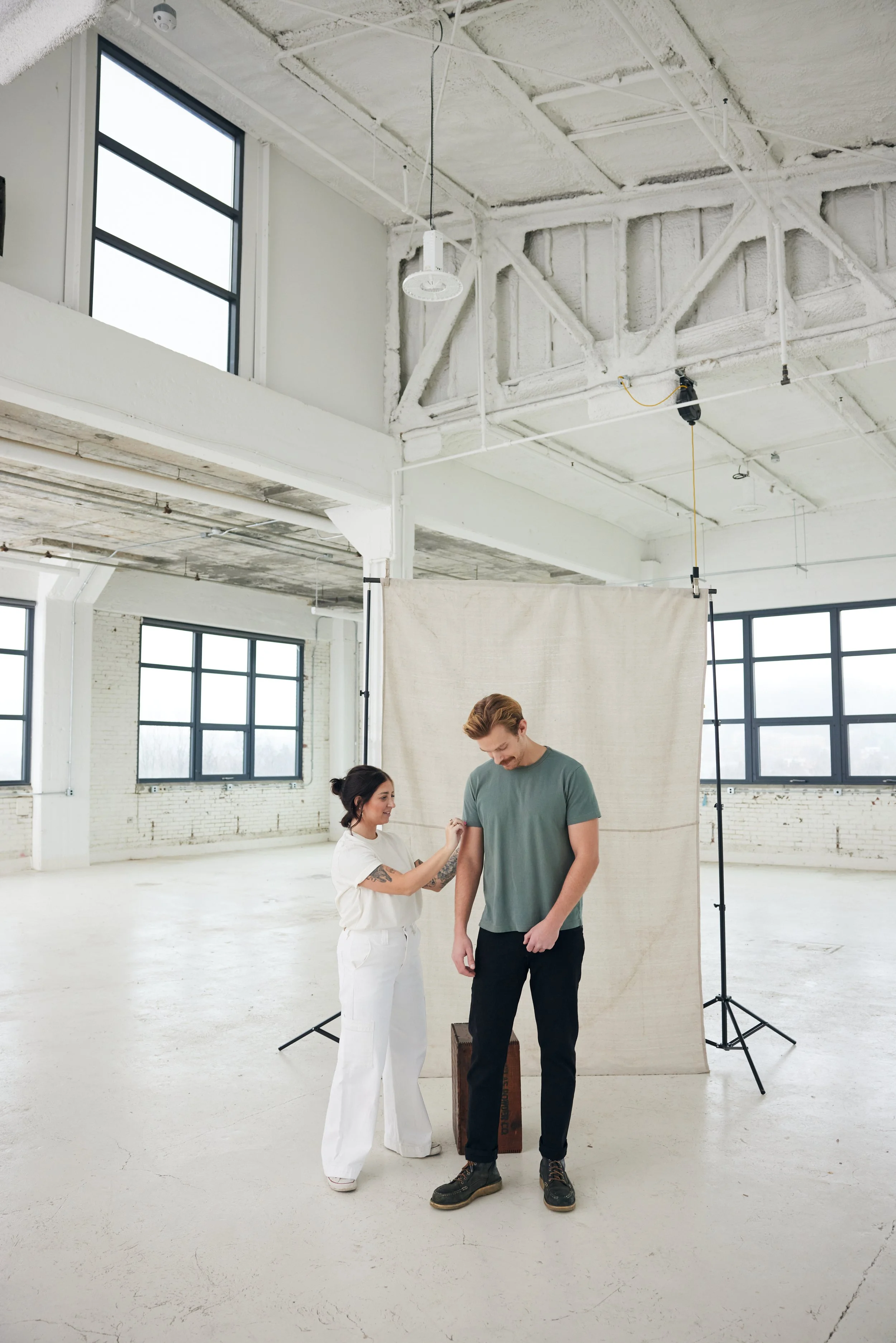 A woman shaves a man's arm in a minimalist, industrial-style photo studio with large windows and a fabric backdrop.
