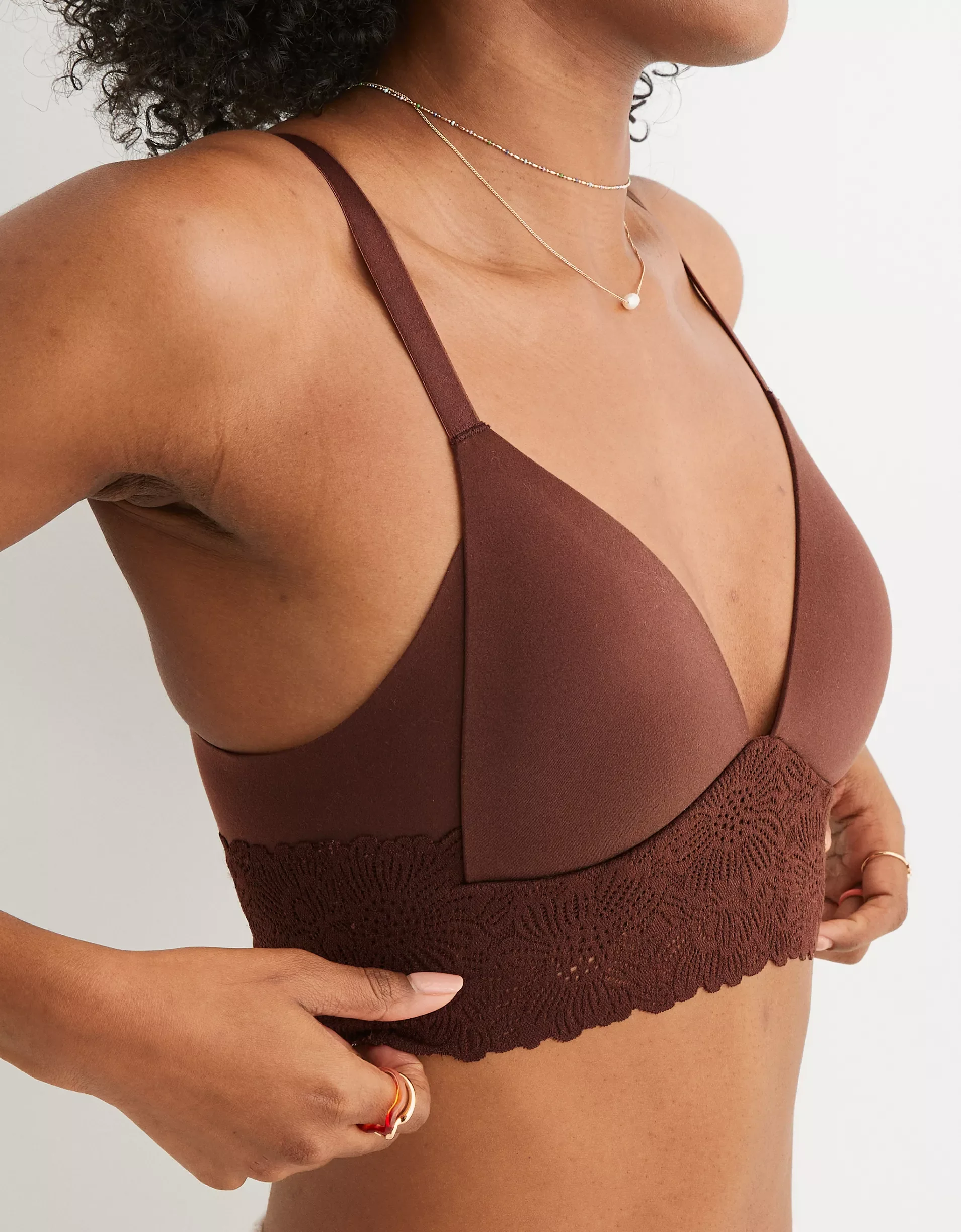 Close-up of a woman wearing a brown bra with lace detail, layered necklaces, and a ring on her finger.