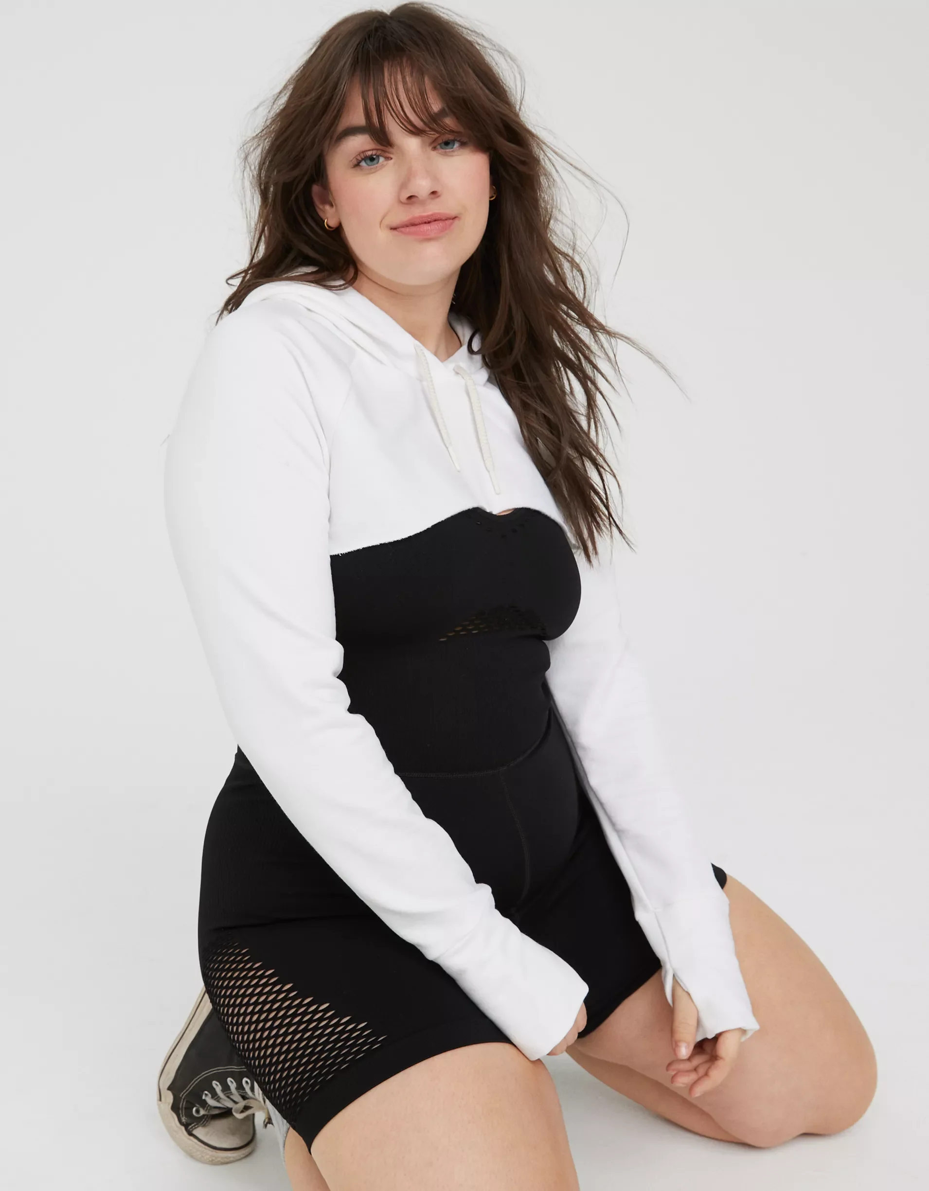 Young woman with brown hair in a white hoodie with black accents, black shorts with mesh panels, and sneakers, kneeling on the floor against a plain white background.