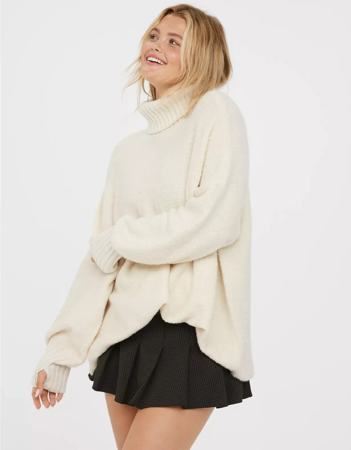 A young woman smiling, wearing a cream turtleneck sweater and a black pleated skirt, standing against a plain white background.