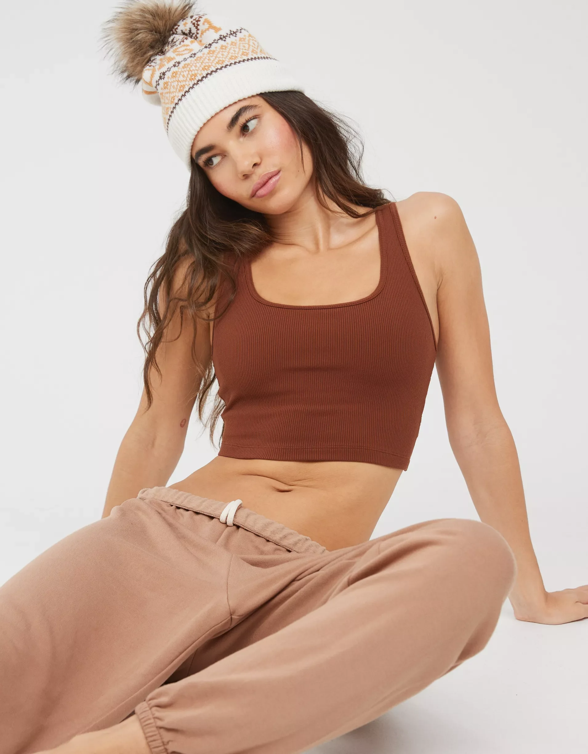 Young woman wearing a white, brown, and orange knitted beanie with a pom-pom, a brown crop top, and beige sweatpants, sitting against a white background.