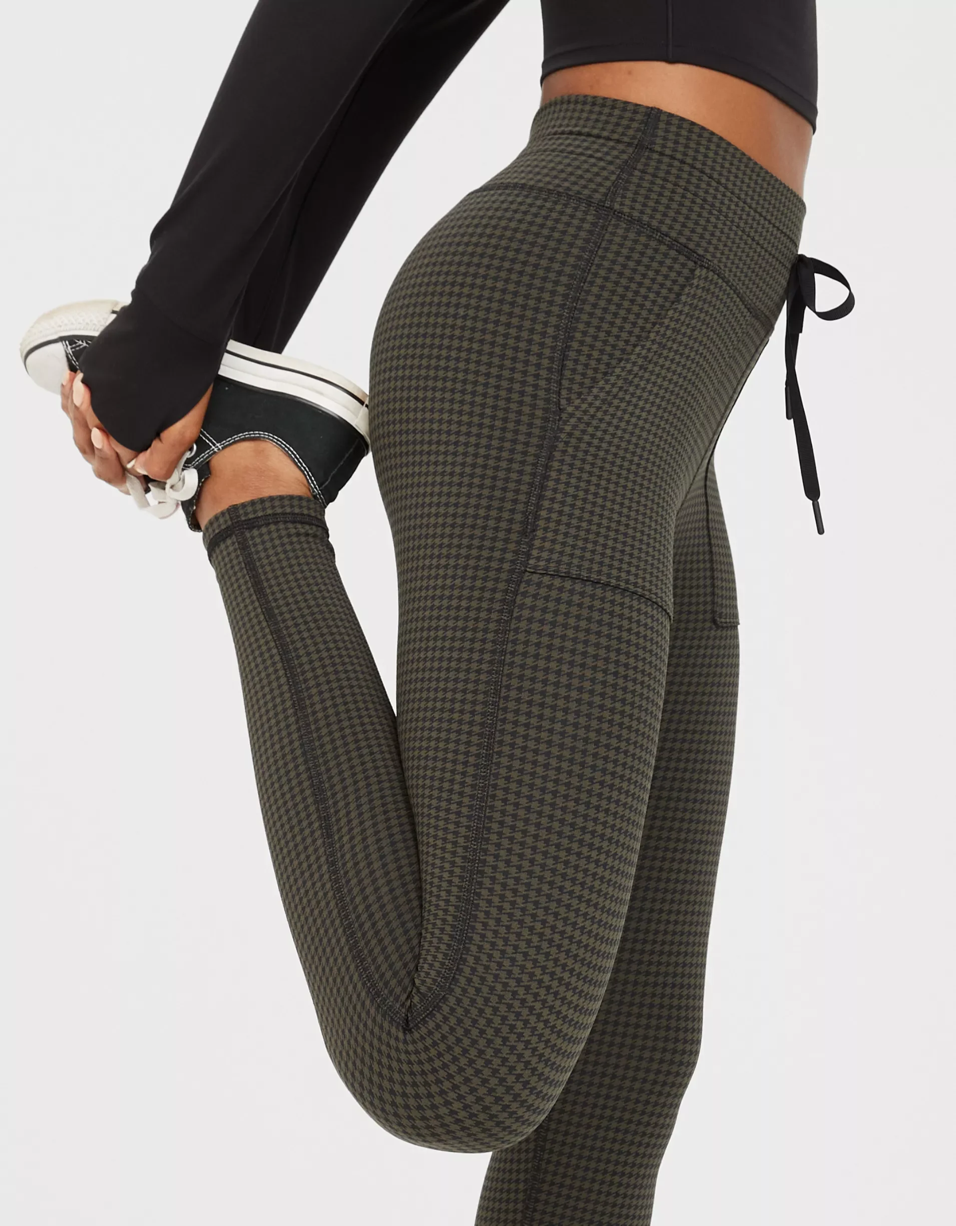 Person in patterned green and black leggings stretching and holding their foot behind them, wearing a black long-sleeve top and sneakers.