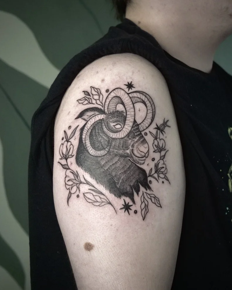 Black and gray tattoo of a ram's head with twisting horns, surrounded by leaves and star-like symbols on a person's upper arm.