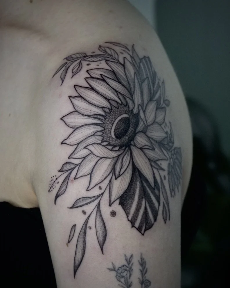 Black and gray sunflower tattoo on person's shoulder with detailed petals and leaves.
