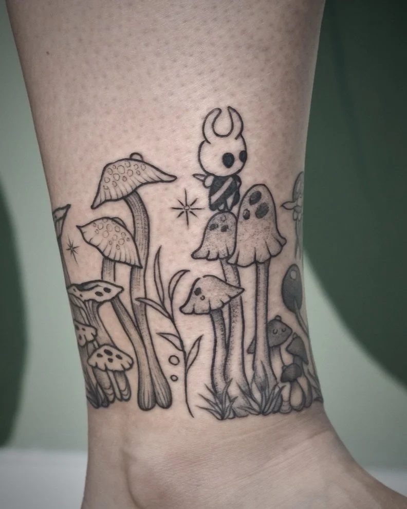 Tattoo of various mushrooms and plants with a small cartoon character with a skull face and horns on top of one mushroom.