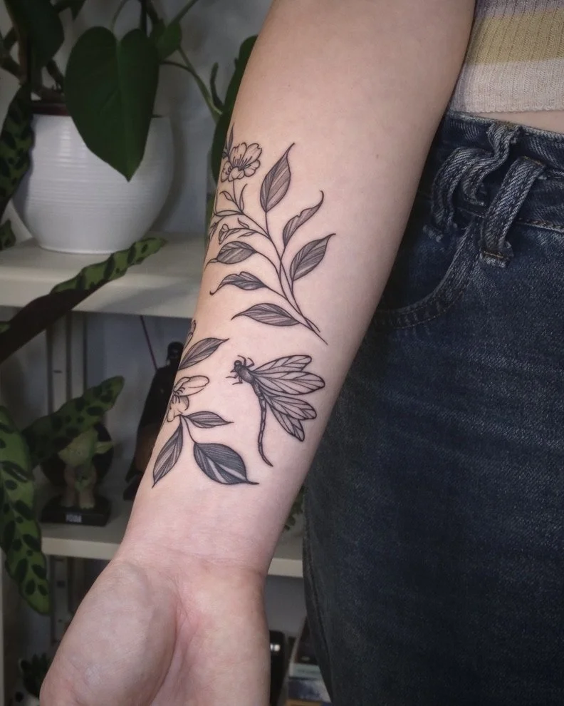 A tattoo of a floral branch with leaves and a dragonfly taken on a person's forearm, with a background of houseplants and indoor furniture.