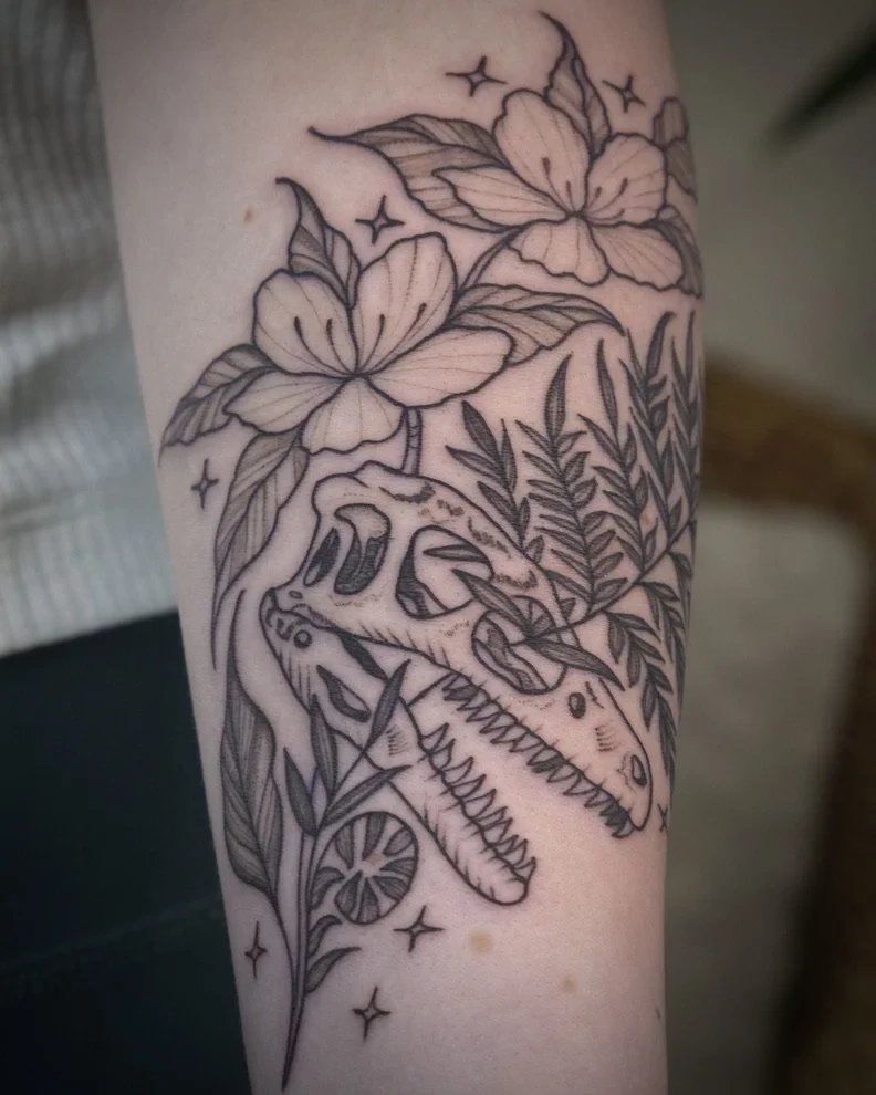 Tattoo of a dinosaur skull surrounded by flowers, leaves, and stars.