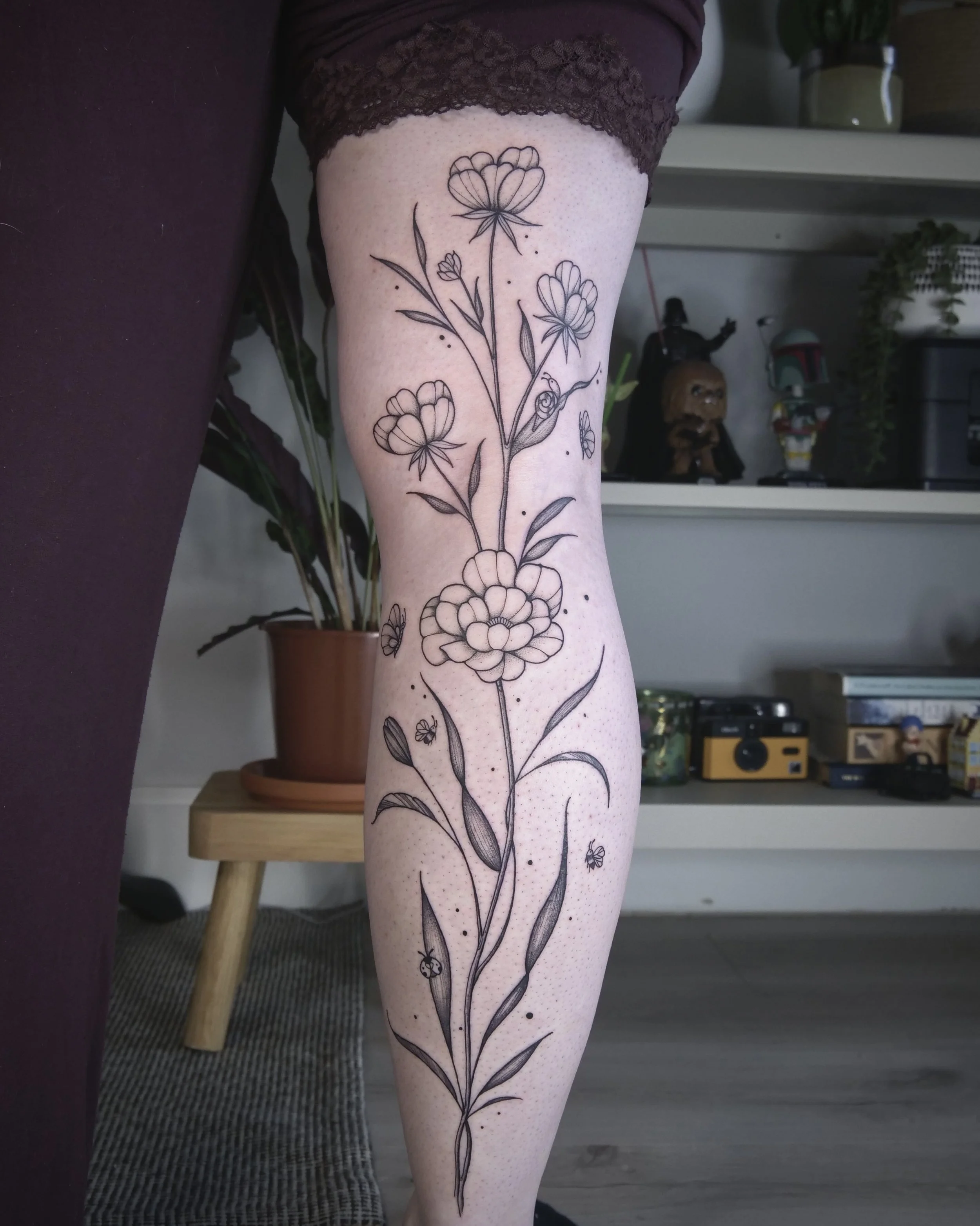 A person's leg with a black line floral tattoo featuring flowers, leaves, and small decorative elements, with a living room background including a wooden table, plant, and shelves.