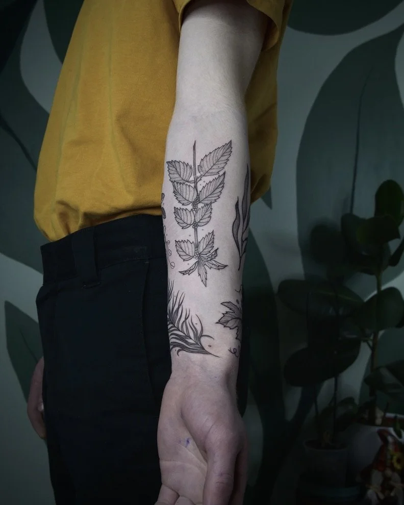 A person wearing a yellow shirt and black pants, showing a tattoo of leaves and flames on their forearm, standing against a wall with leaf patterns.