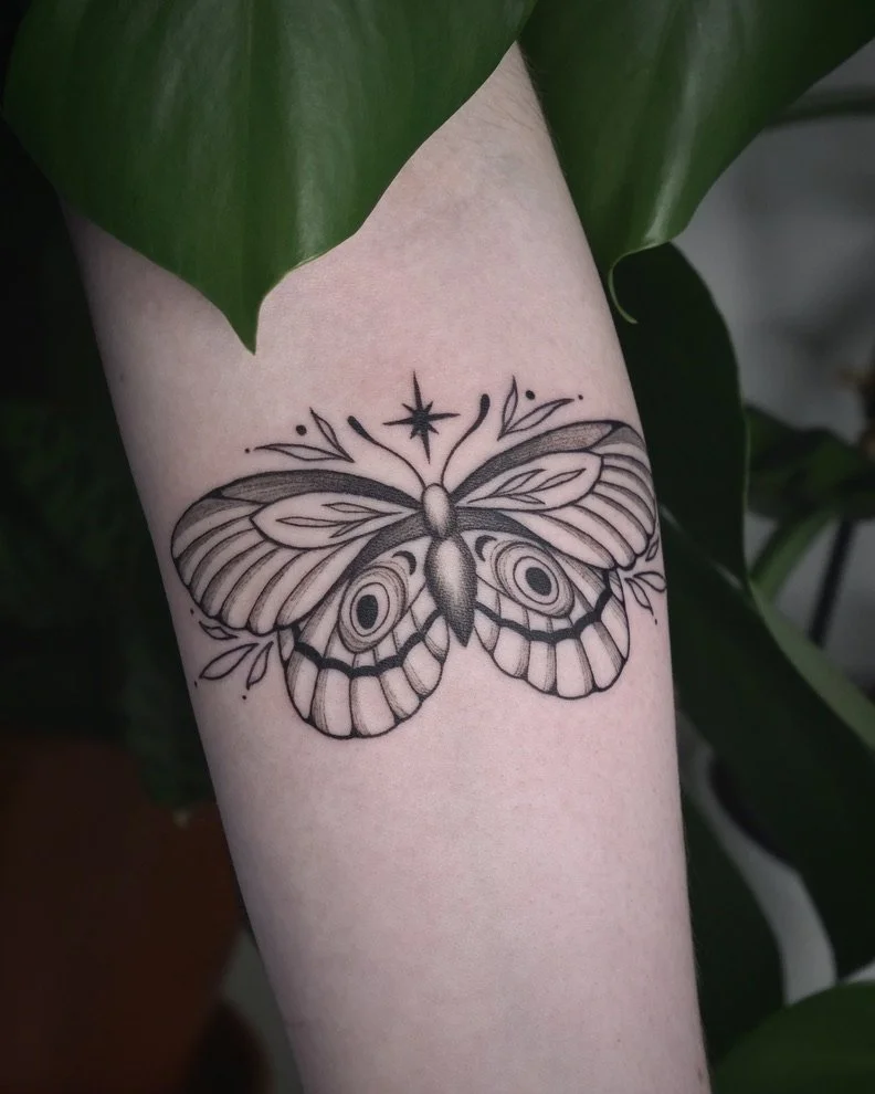 A black and gray tattoo of a detailed butterfly with a star above it, surrounded by leaves on the arm.