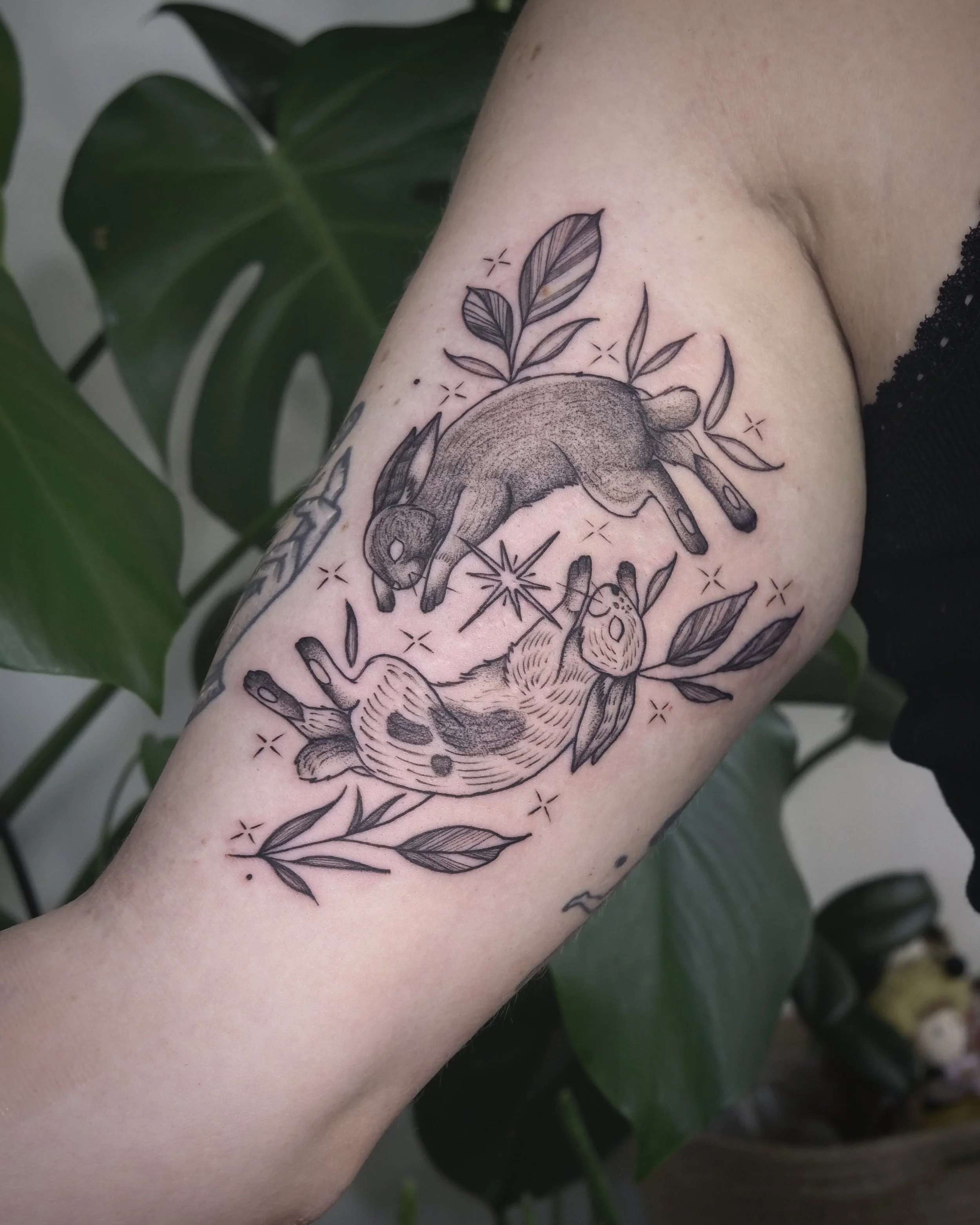 Line art tattoo of a sleeping fox and a curled-up dog surrounded by leaves and stars on a person's upper arm.