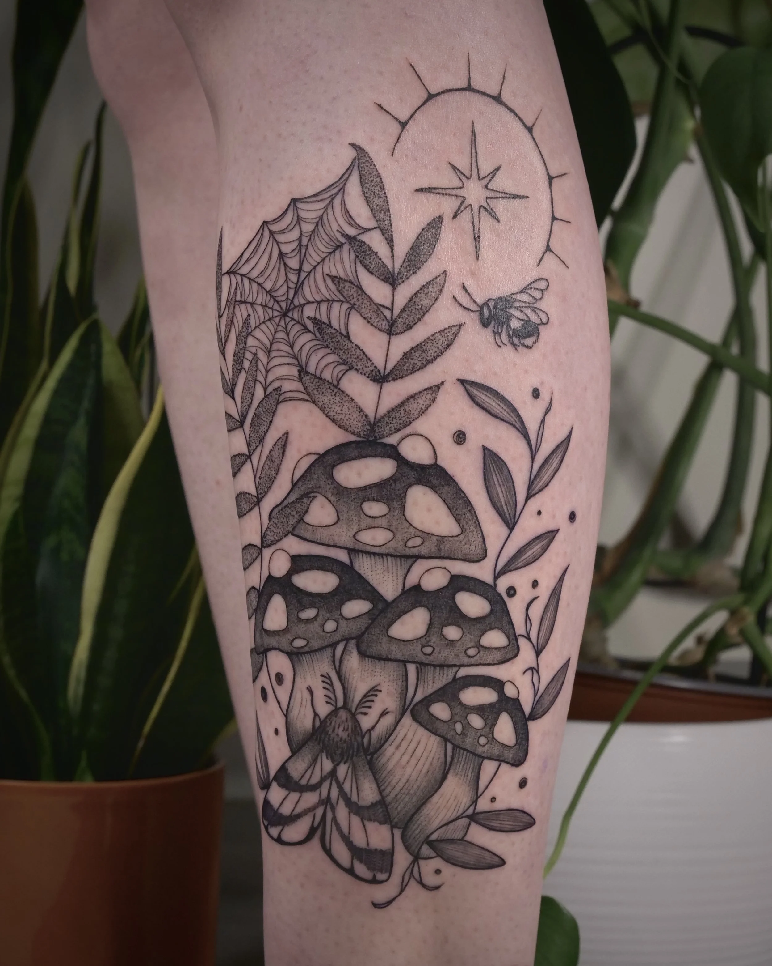 Tattoo of fungi, leaves, a spider, a bee, and a sun or star on a person's leg.