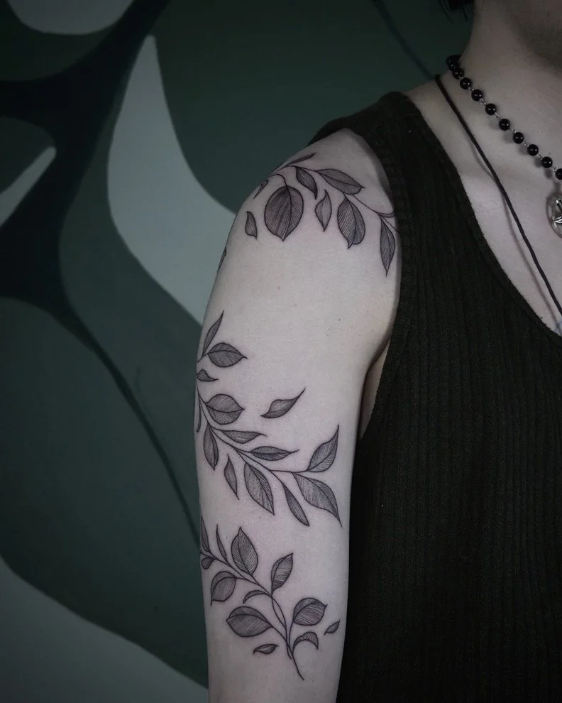 Black ink tattoo of a leafy vine on person's upper arm, with leaves extending from shoulder to elbow, against a dark abstract background.
