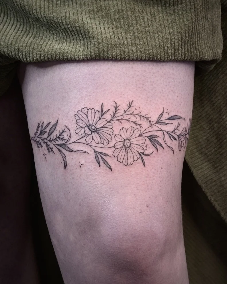 Black and grey floral tattoo with daisy-like flowers and leaves on a person's thigh.