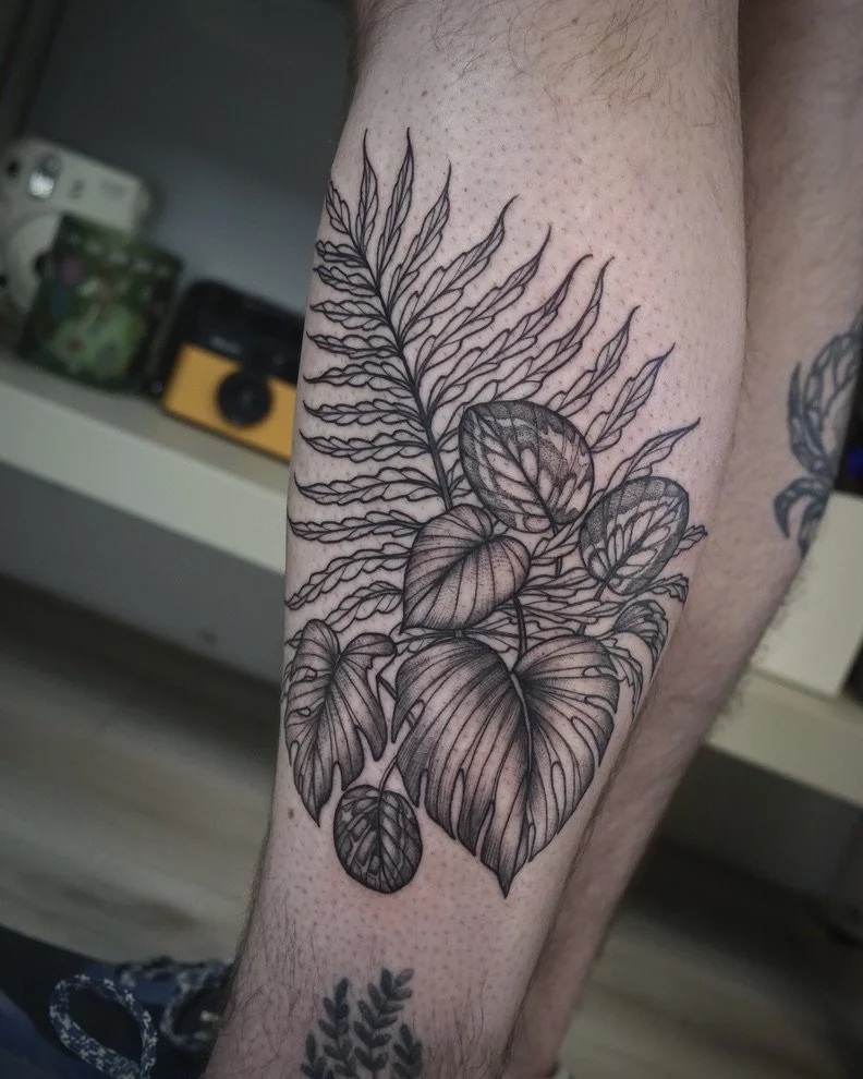 Black and gray botanical tattoo of a fern frond and various leaves on a person's forearm.
