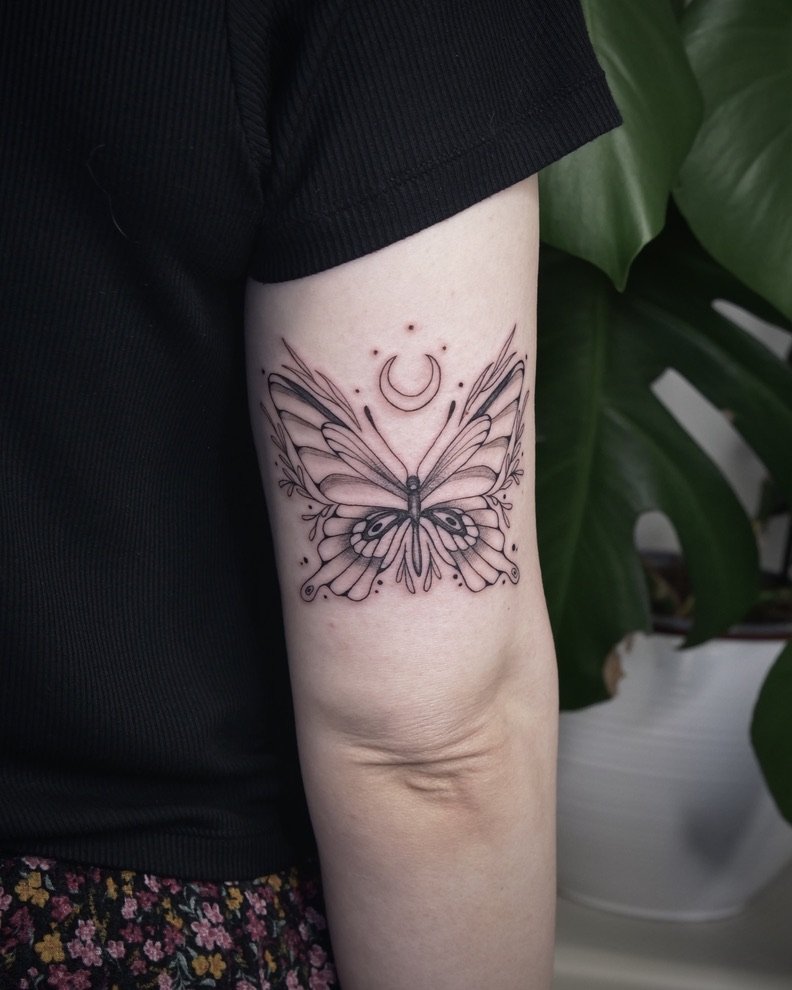 A tattoo of a butterfly with detailed wings, a crescent moon, and small celestial dots on the person's upper arm. Part of the person's black shirt and floral-patterned skirt are visible, with a large green plant in the background.