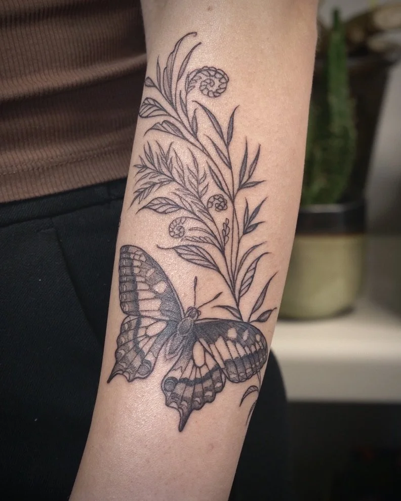 Black outline tattoo of a butterfly perched on a plant with leaves, berries, and flowers on a person's forearm.