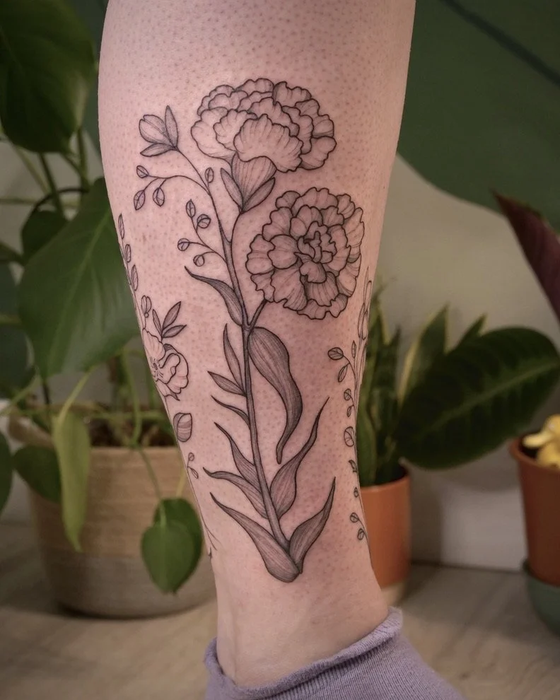 A black and gray outline tattoo of two large flowers with detailed petals, along with leaves and smaller buds, on a person's lower leg.