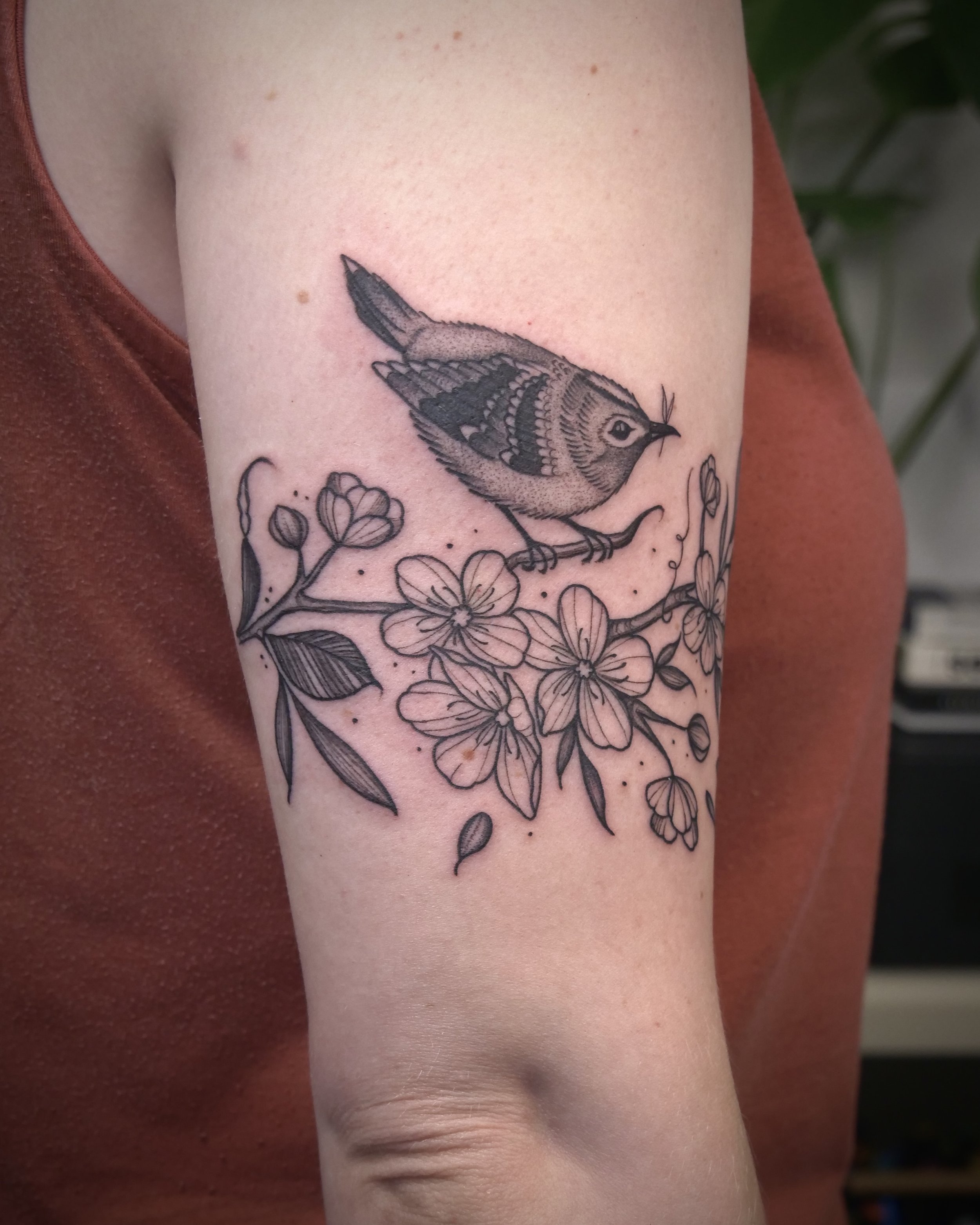 Black and gray tattoo of a small bird perched on a branch with flowers on the person's upper arm.