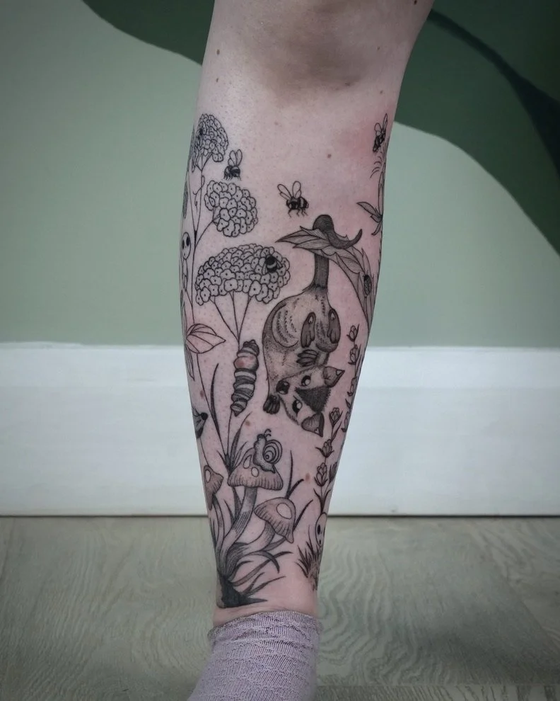 Black ink tattoo of a forest scene with mushrooms, a squirrel, insects, and plants on a person's lower leg.