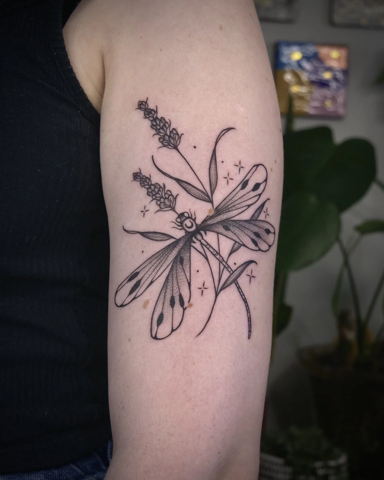 Line art tattoo of a dragonfly with detailed wings and bouquet of lavender flowers on person's upper arm.