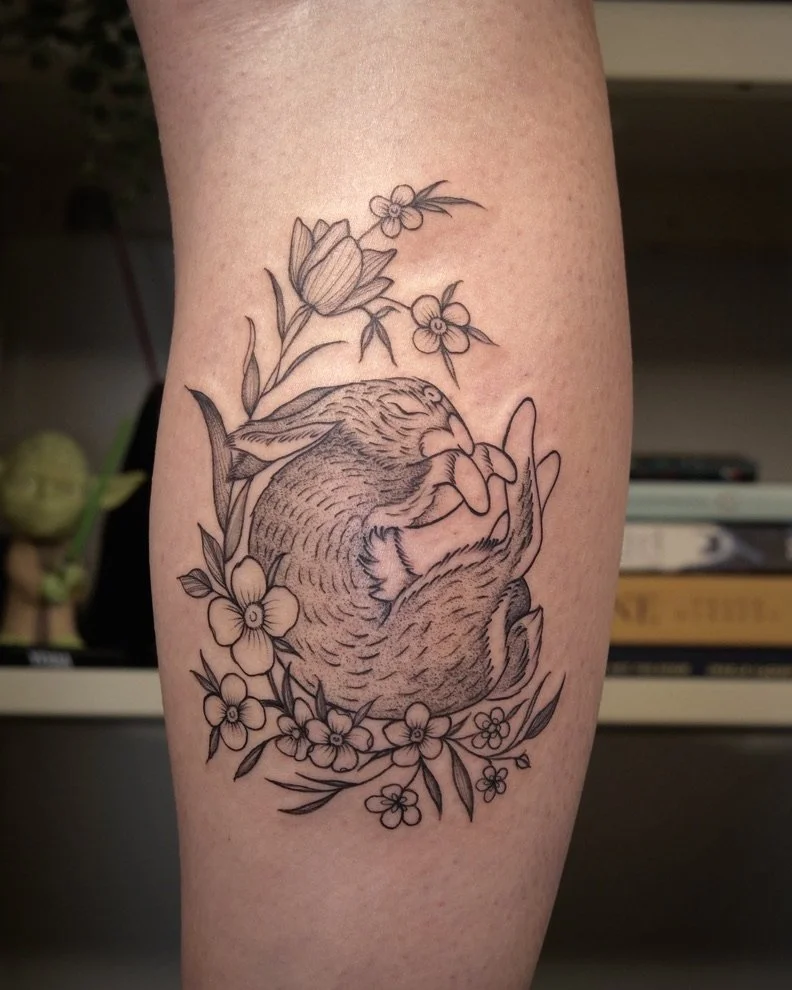 Black outline tattoo of a sleeping fox curled up among flowers and leaves on a person's arm.