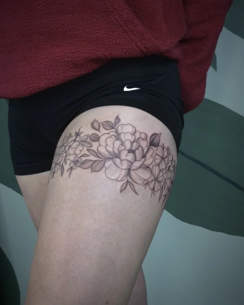 A person's upper thigh with a large, detailed black outline floral tattoo of various flowers and leaves.