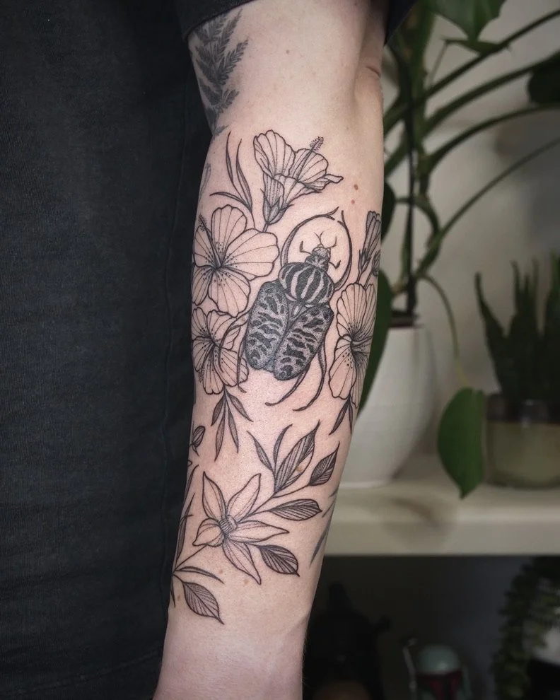 Tattoo of flowers and a moth on person's arm, with plants in background.