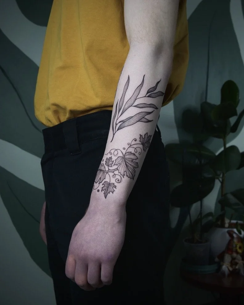 A person's forearm with a large black ink tattoo of leaves and vines.