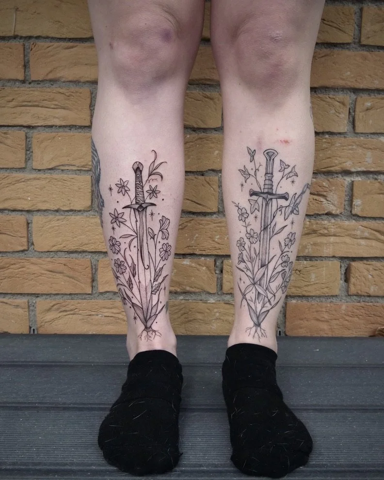 Two legs with floral and sword tattoos on the shins, standing against a brick wall.