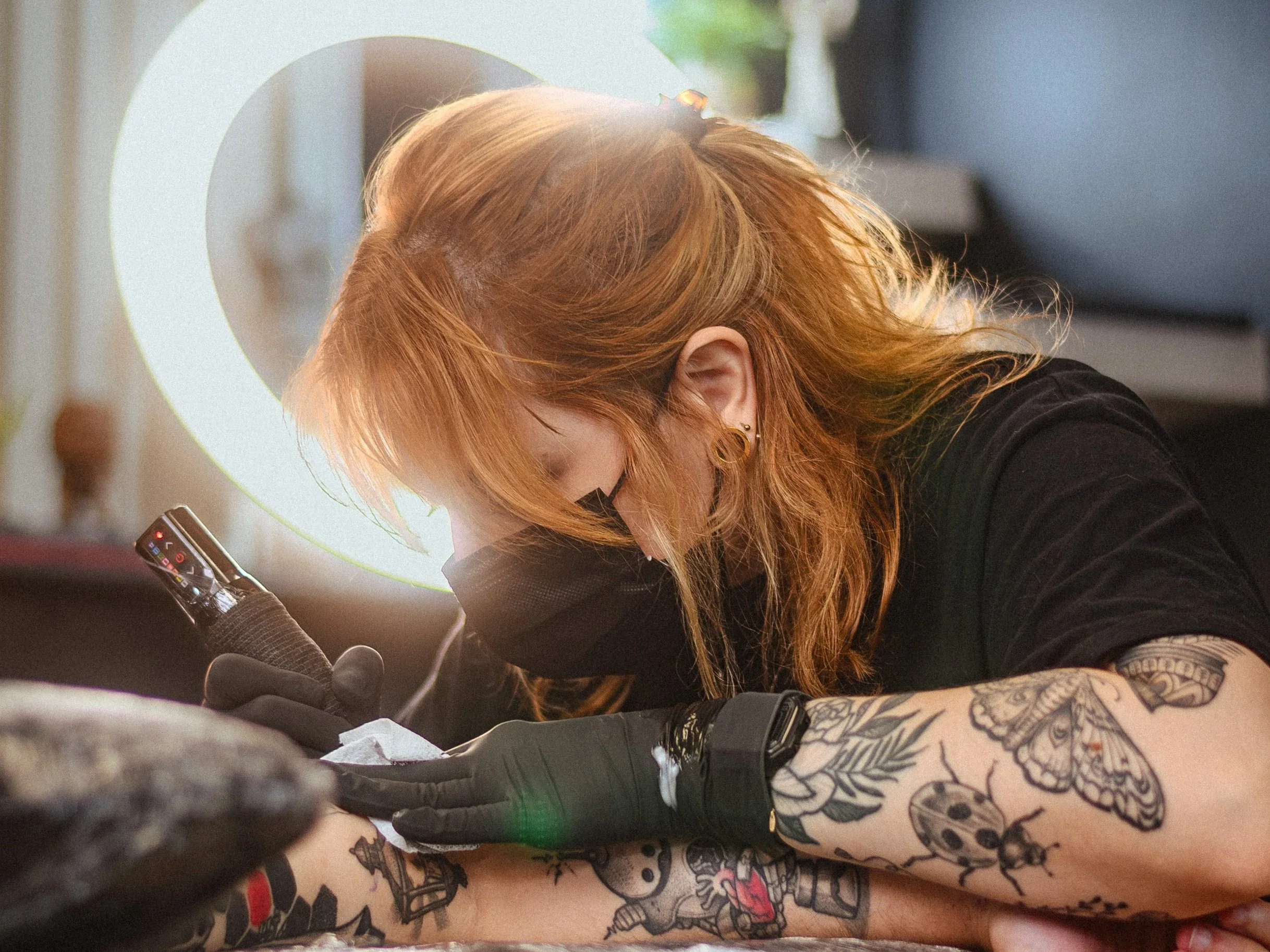 A tattoo artist with orange hair, glasses, and tattoos on both arms is working on a tattoo while wearing a black mask, gloves, and a black shirt. She is focused on her work with a tattoo machine in hand, with a vanity mirror and blurred background in the setting.