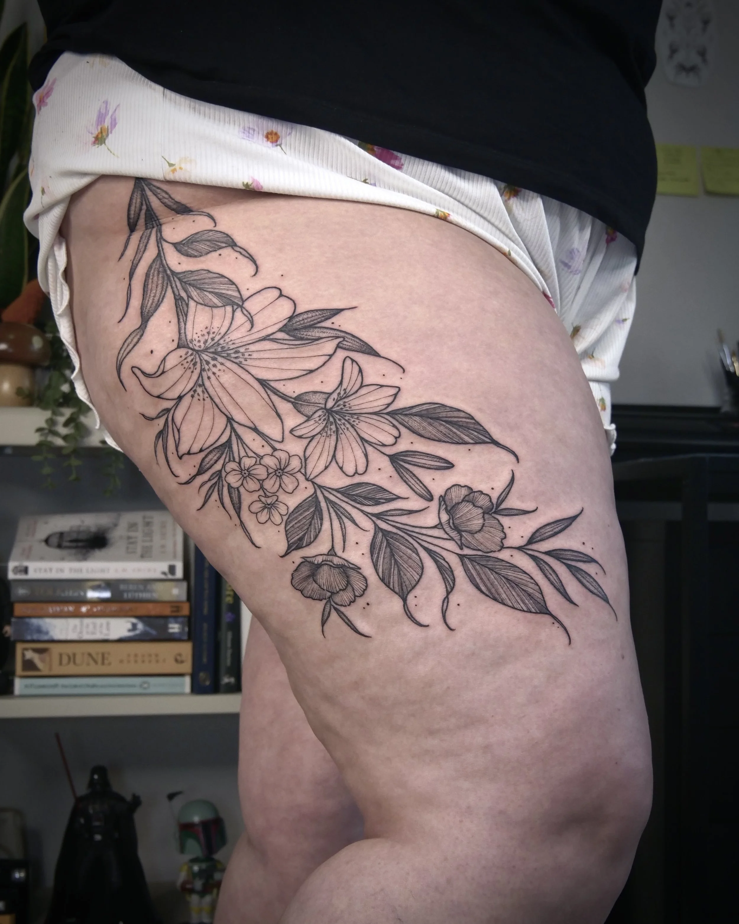 A black outline tattoo of a floral design on a person's upper thigh, featuring large lilies, smaller flowers, and leaves.