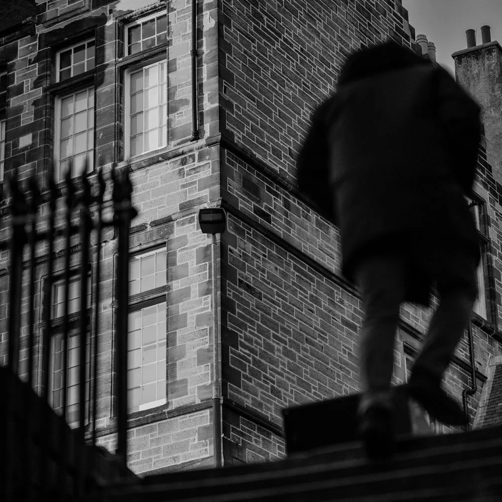 Black and white square street photograph of a solitary figure walking up stone steps between urban architecture, captured in a minimalist contemporary documentary style.