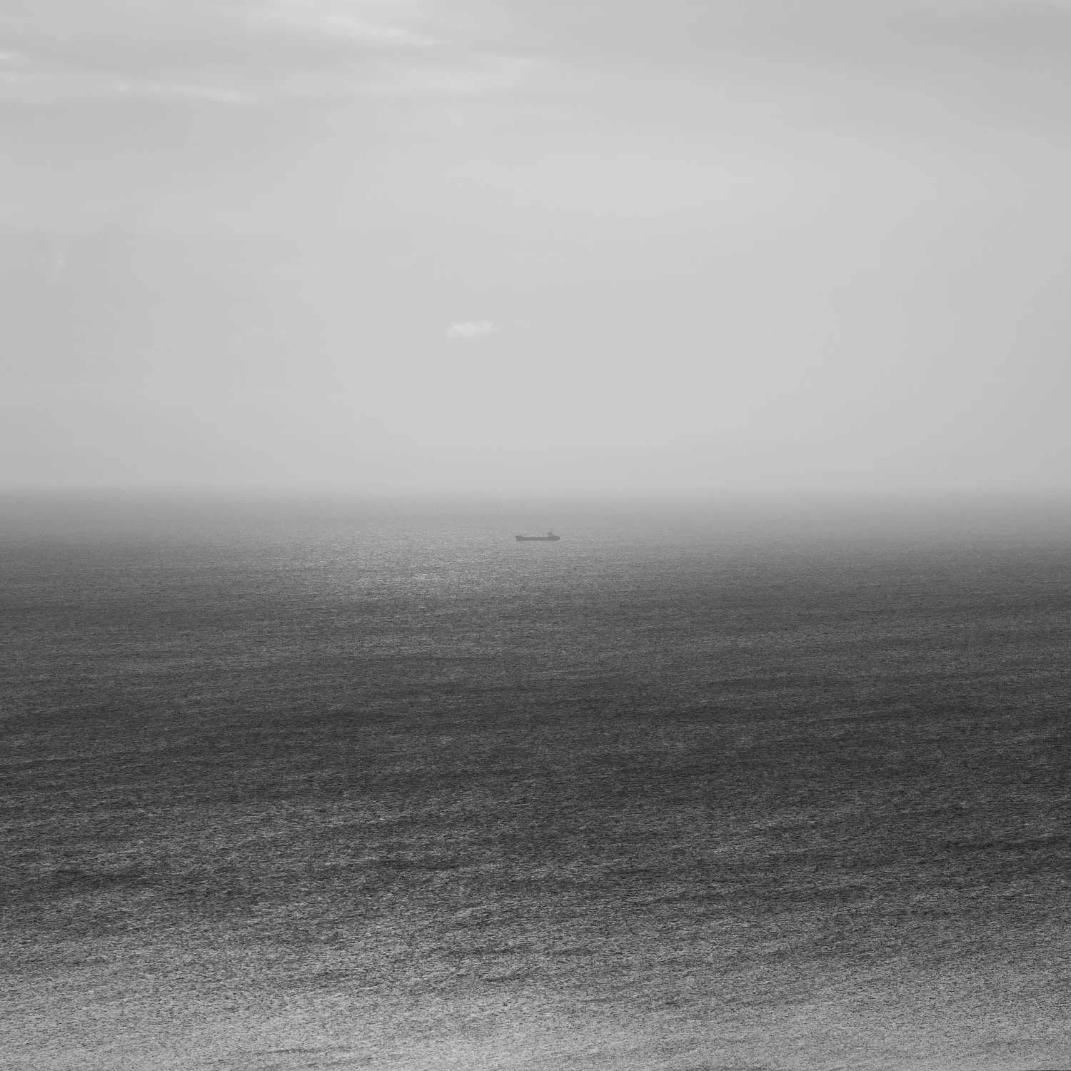 Black and white minimalist seascape photograph showing a lone cargo ship on the horizon, surrounded by misty sky and textured sea surface.