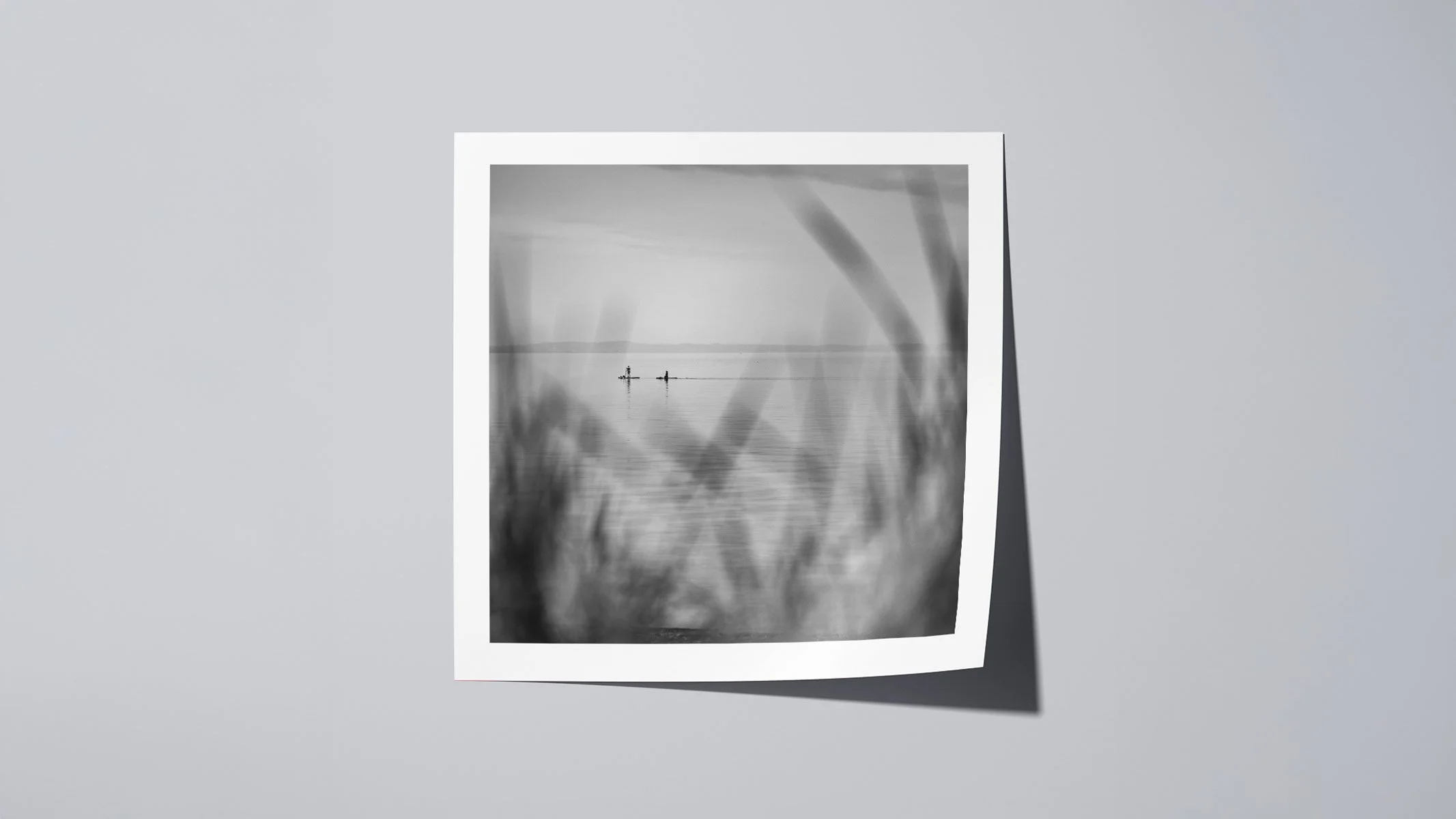 Black and white fine art photography print presented on a neutral background, calm coastal scene with distant paddleboarders.