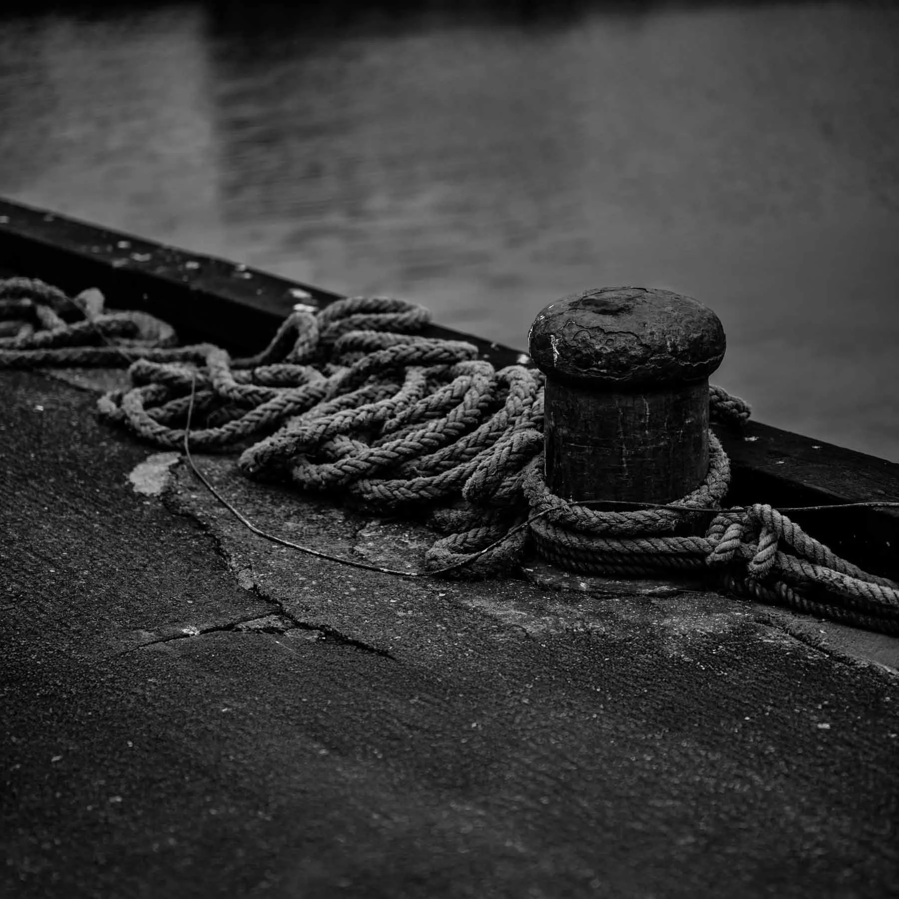 Black and white square photo of a mooring bollard with coiled rope on weathered concrete, emphasizing texture, form, and industrial simplicity by the water’s edge.