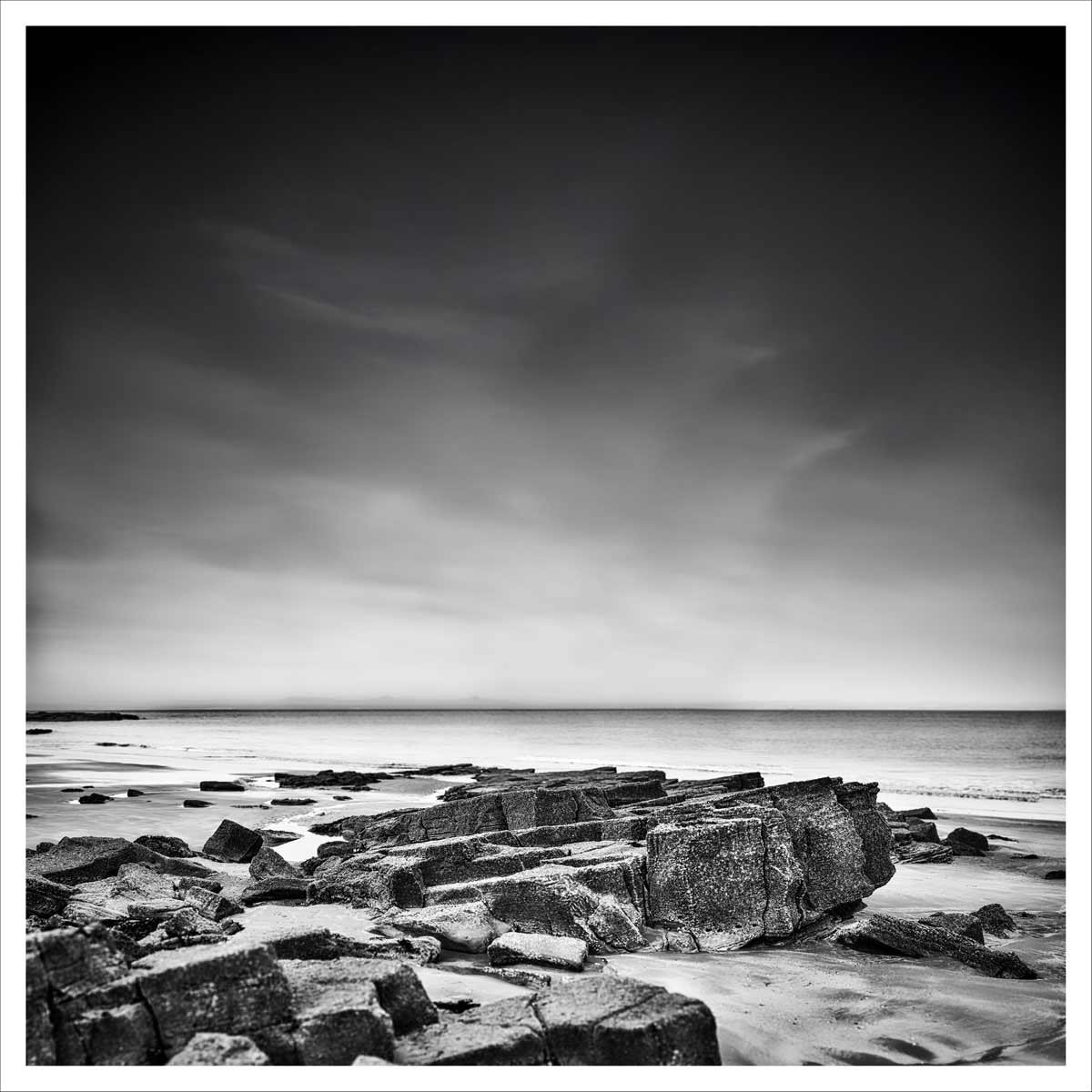 Black and white photograph of a solid rock formation on a quiet shoreline, captured in soft long exposure light.