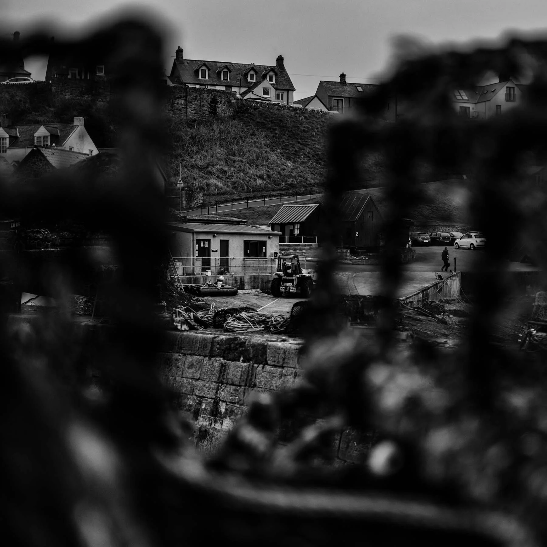 A candid black and white image of a coastal village, partially obscured by lobster creels in the foreground—adding layered depth and mystery.