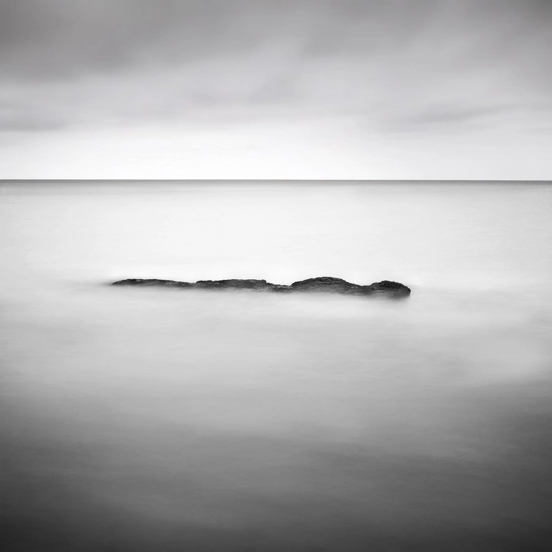 Black and white photo of a rock in calm water under a cloudy sky.
