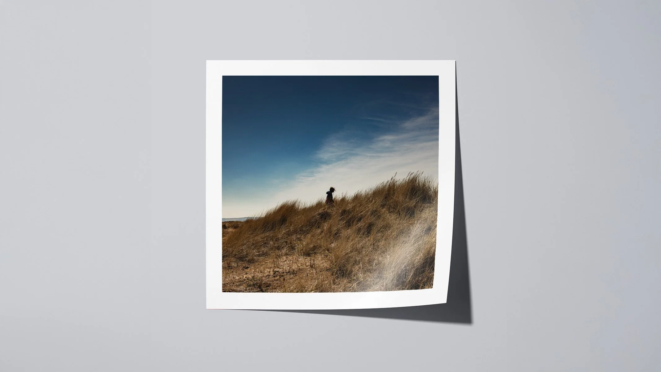 Square fine art photograph of a child exploring grassy sand dunes near the sea on Kinshaldy Beach, Scotland.